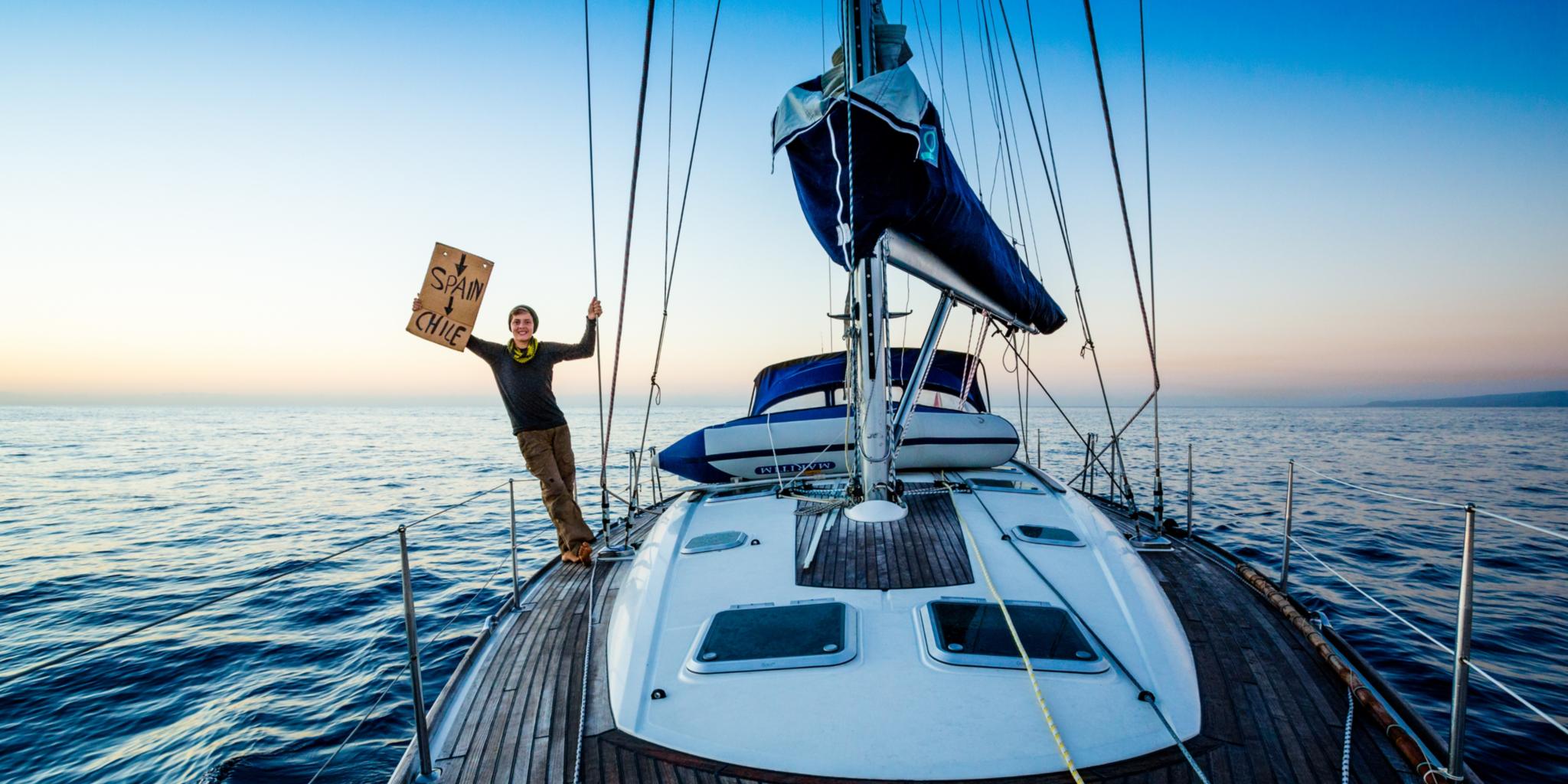 Joshi Nichell steht auf einem weißen Segelboot, das sich auf offener See befindet. Mit einer Hand hält er sich am Segel fest und lehnt sich über das Boot hinaus; mit der anderen Hand hält er ein Schild fest.