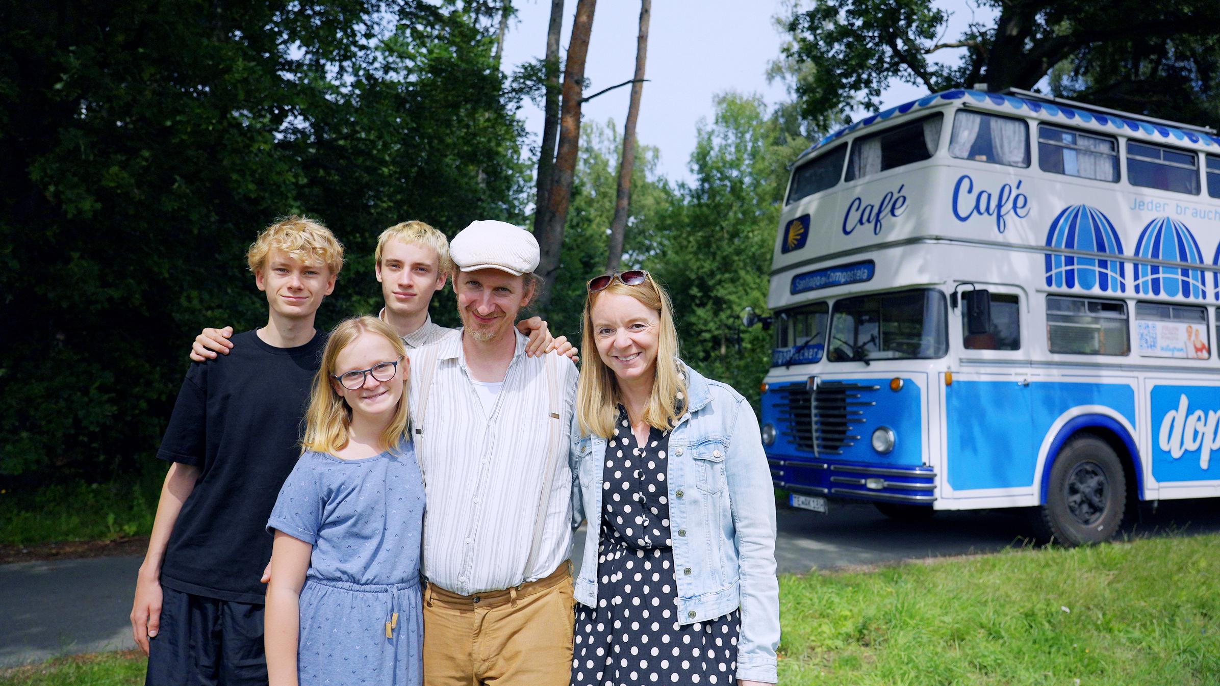 4 Menschen lächeln in die Kamera, hinter ihnen ein großer blau-weißer Bus