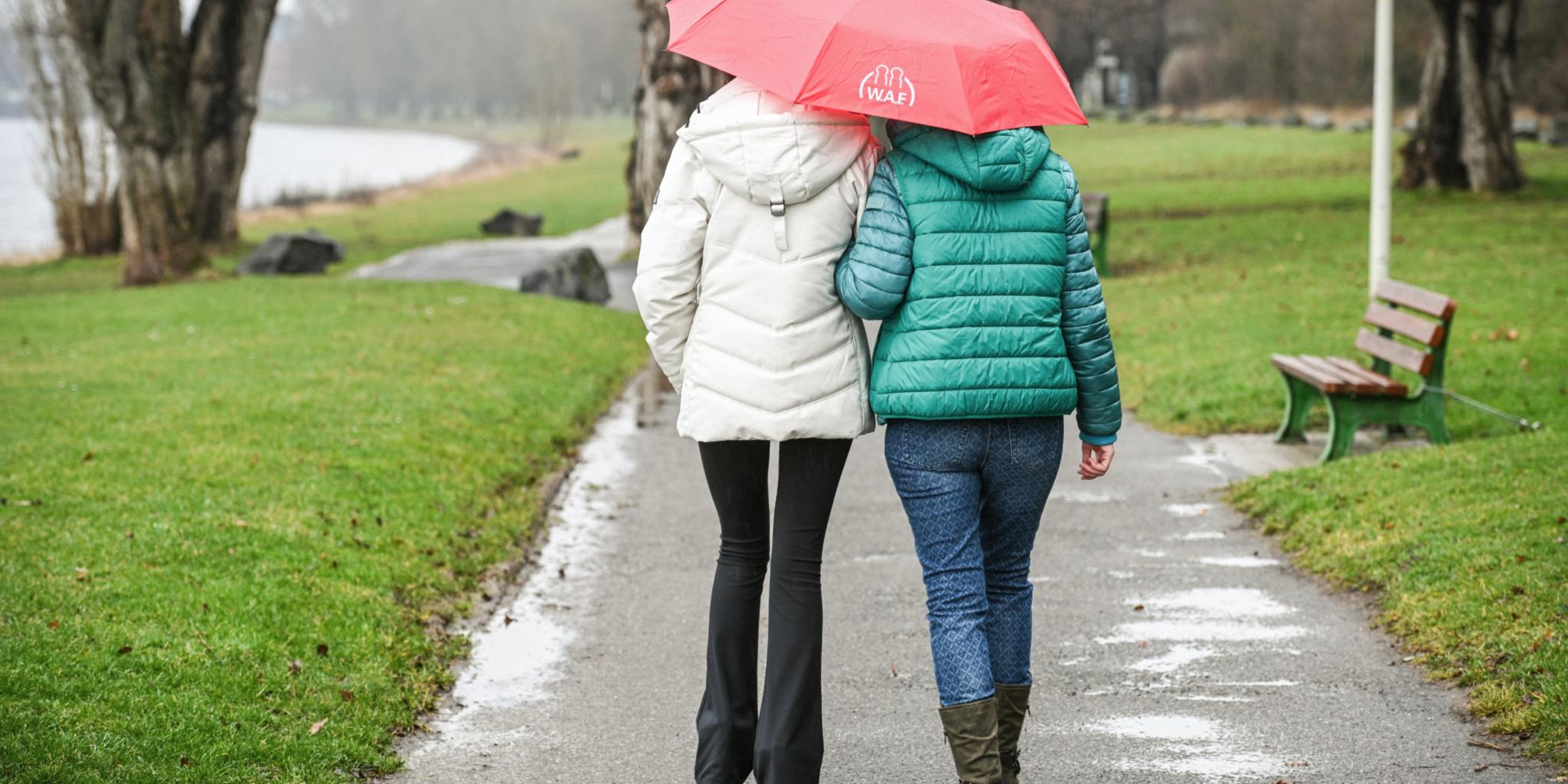 Zwei Frauen aus der Rückansicht spazieren mit einem roten Regenschirm einen Weg entlang