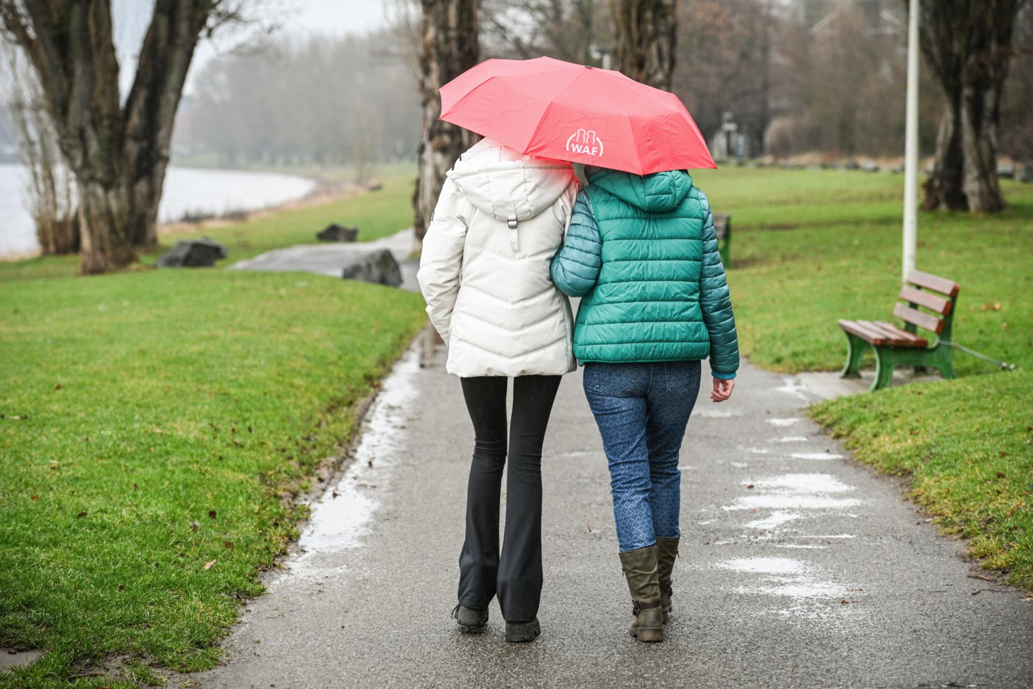 Zwei Frauen aus der Rückansicht spazieren mit einem roten Regenschirm einen Weg entlang