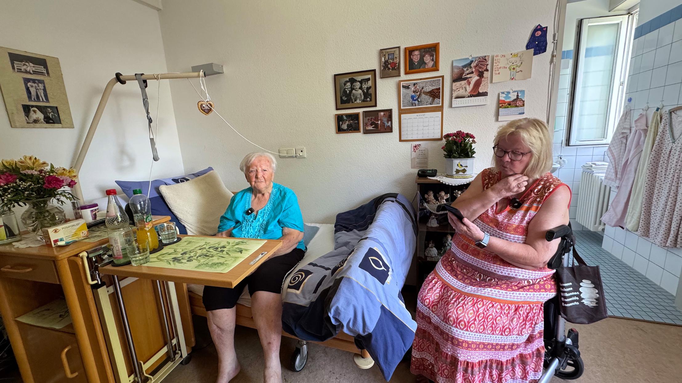 Eine frau sitzt auf dem Rand eines Pflegebettes, daneben sitzt eine Frau auf einem Rollator