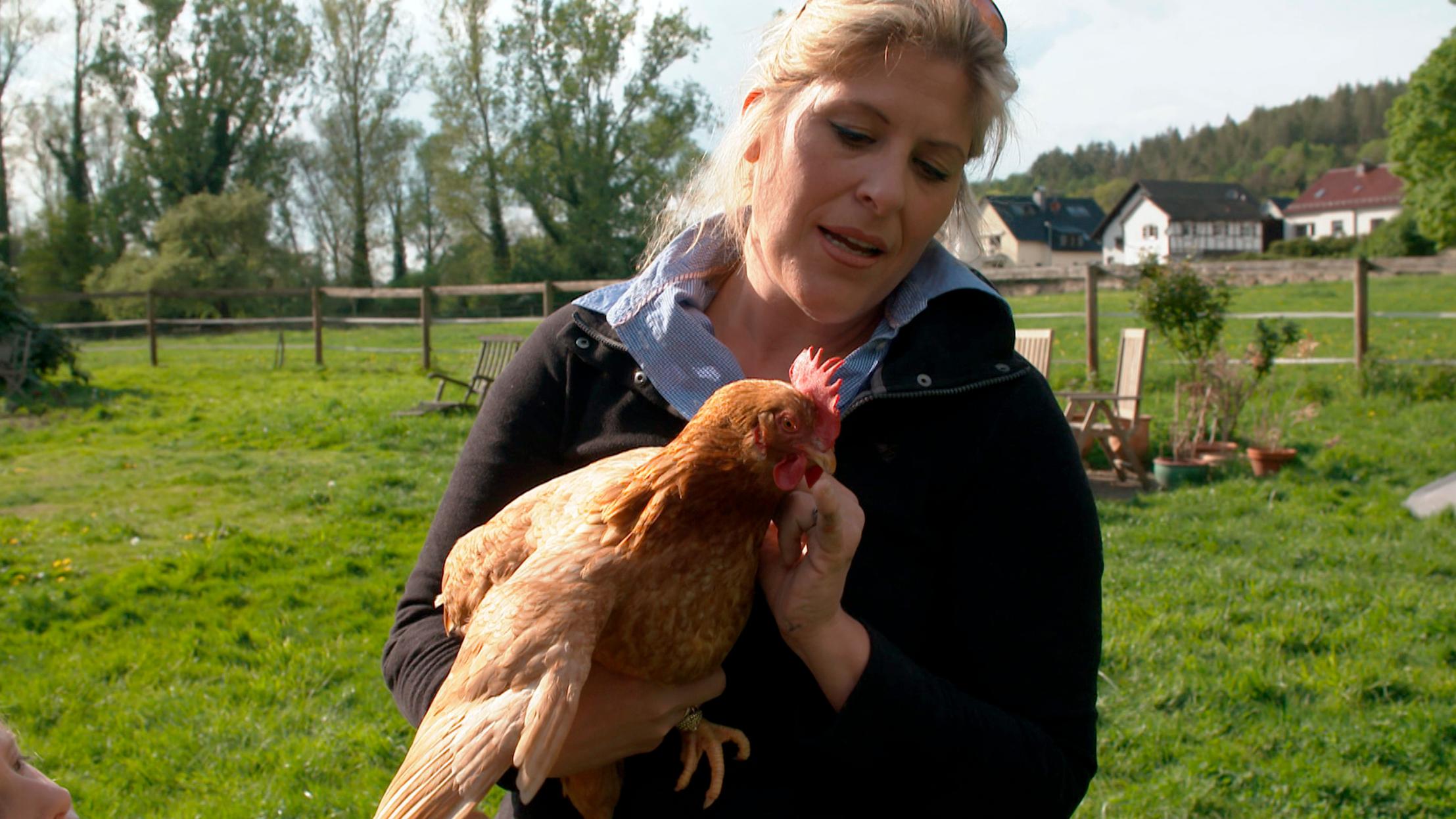 Eine blonde Frau hält ein braunes Huhn auf dem Arm und steht auf einer grünen Wiese