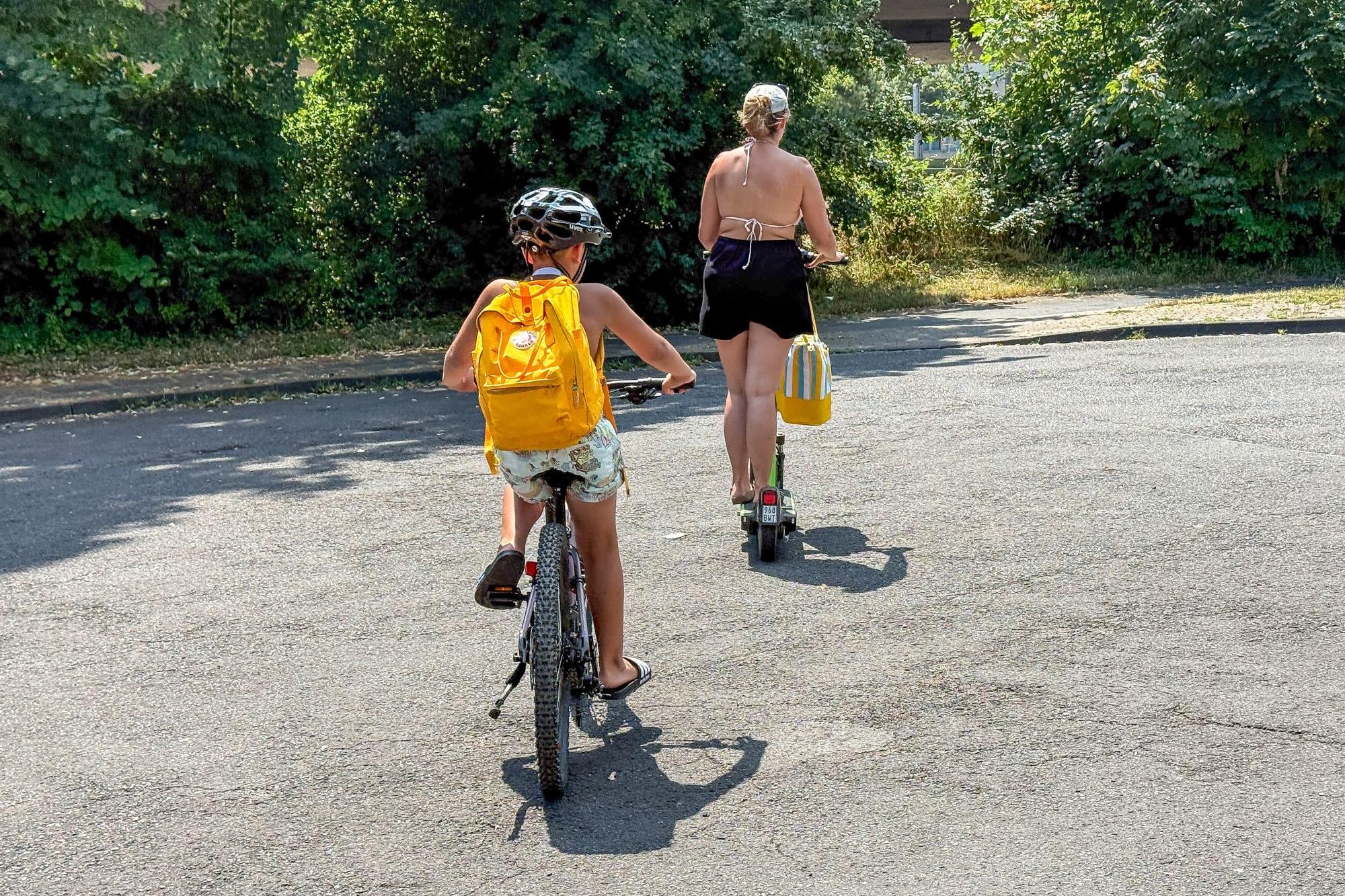 Ein Kind mir gelbem Rucksack fährt Fahrrad, davor geht eine Frau