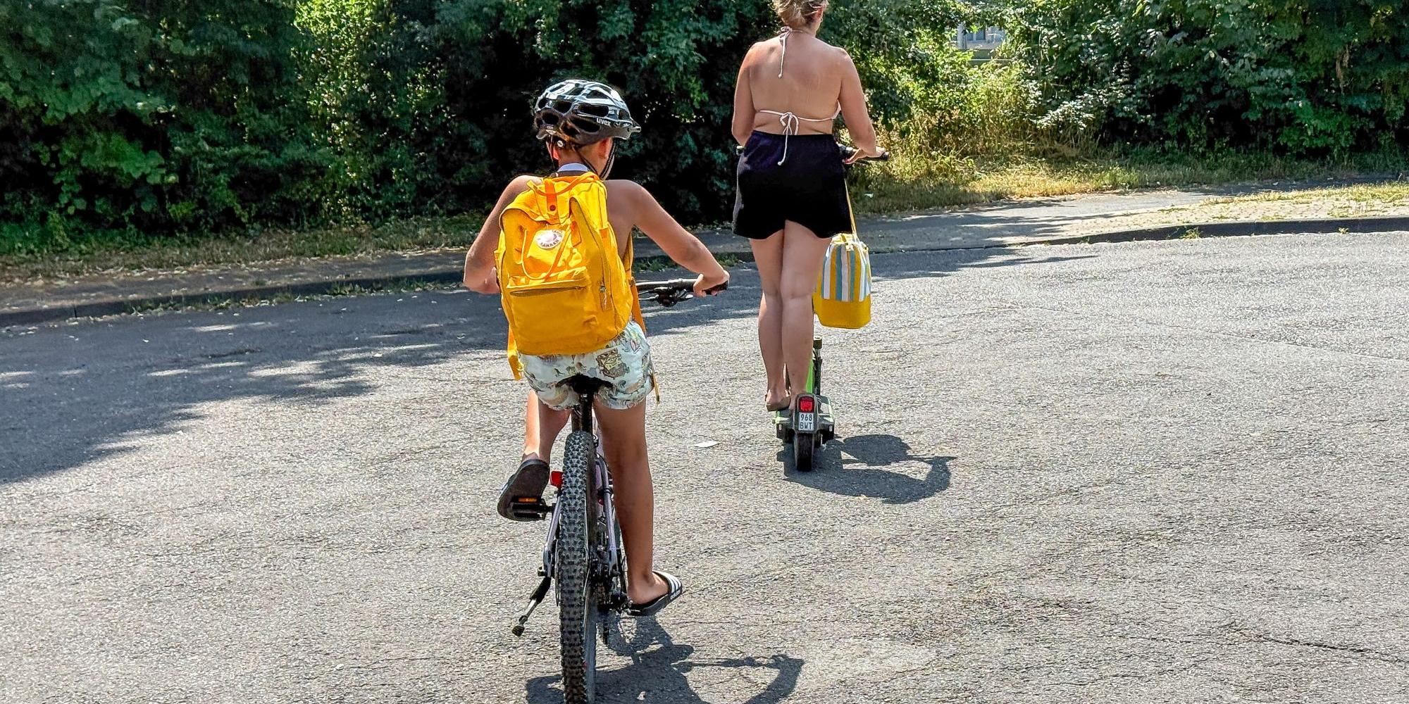 Ein Kind mir gelbem Rucksack fährt Fahrrad, davor geht eine Frau
