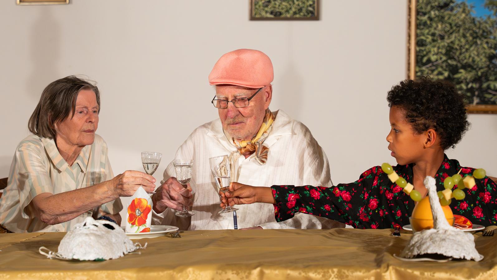 Szene eines Theaterstücks. Zwei Senioren und ein Kind stoßen mit ihren Gläsern an.