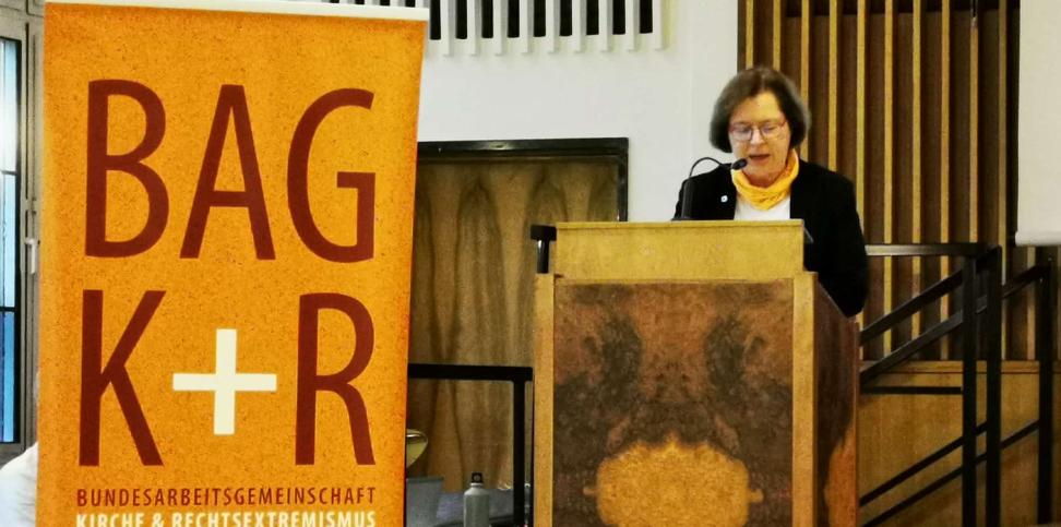 Eine Frau steht an einem Rednerpult, daneben ein Roll-Up mit dem BAG K+R Logo