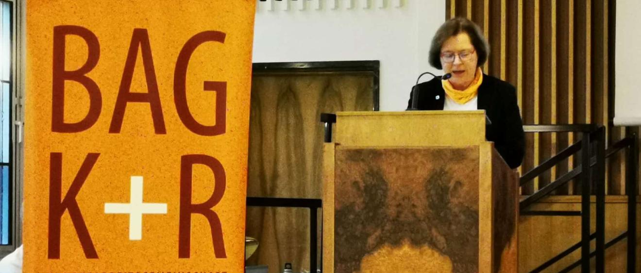 Eine Frau steht an einem Rednerpult, daneben ein Roll-Up mit dem BAG K+R Logo