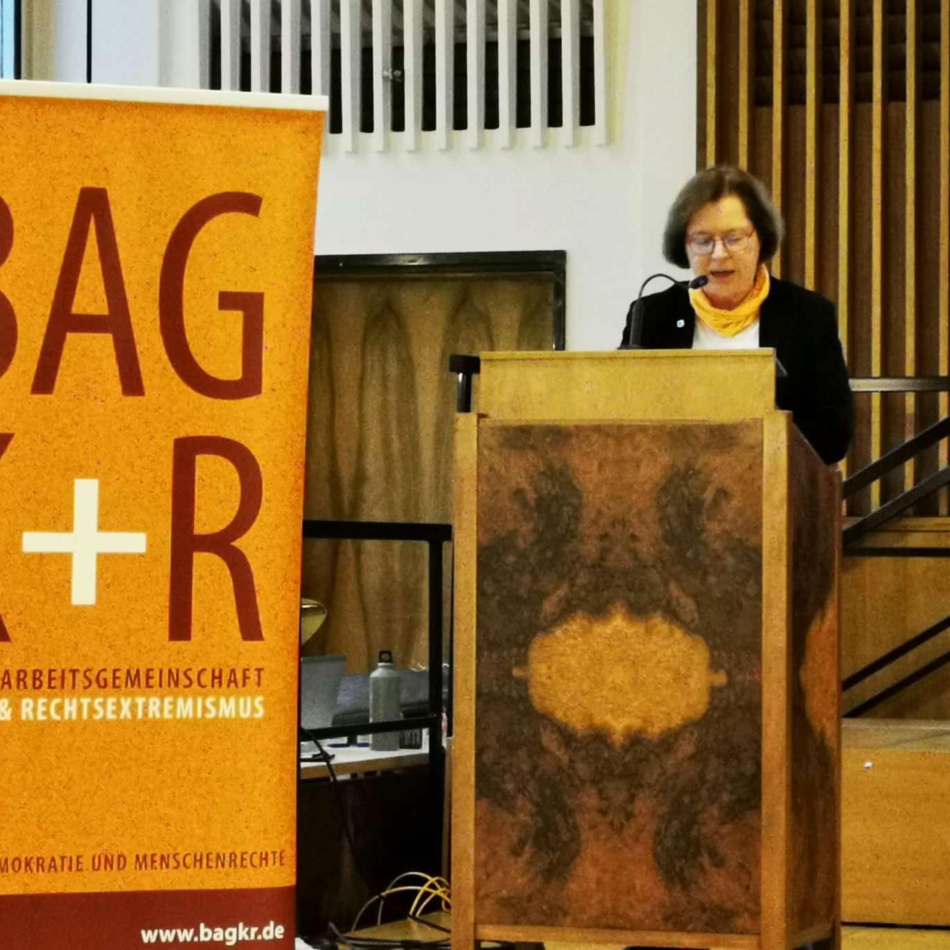 Eine Frau steht an einem Rednerpult, daneben ein Roll-Up mit dem BAG K+R Logo