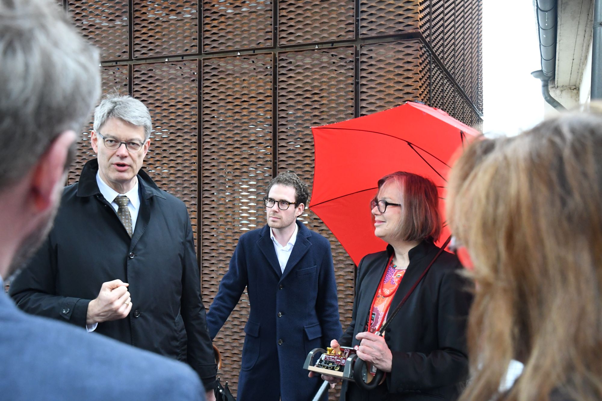Ein Mann und eie Frau mit einem roten Regenschirm sprechen mit Menschen an einem Bahnhof