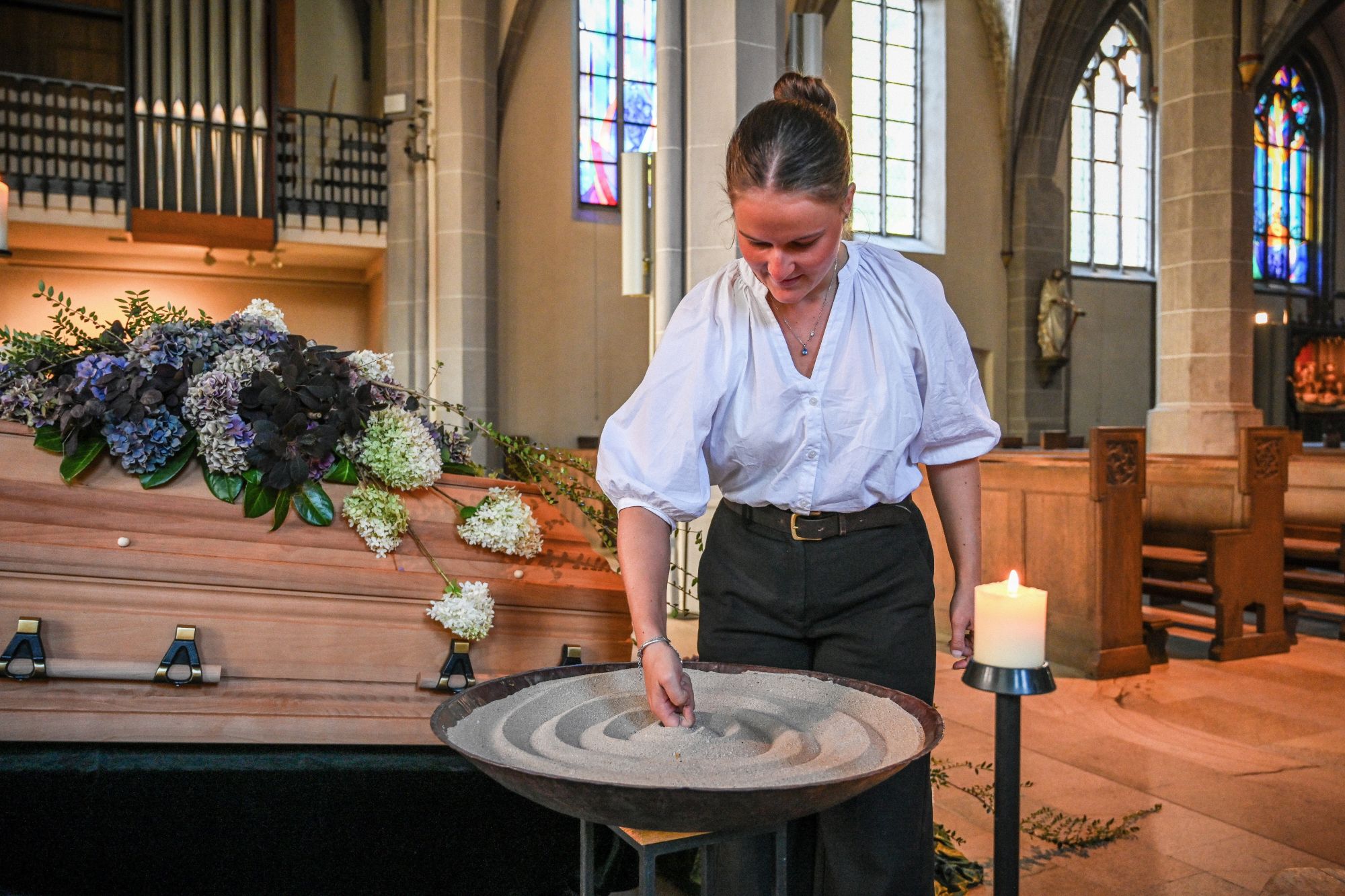 Eine Bestatterin gestaltet Sand in einem Gefäß vor einem geschmückten Sarg.