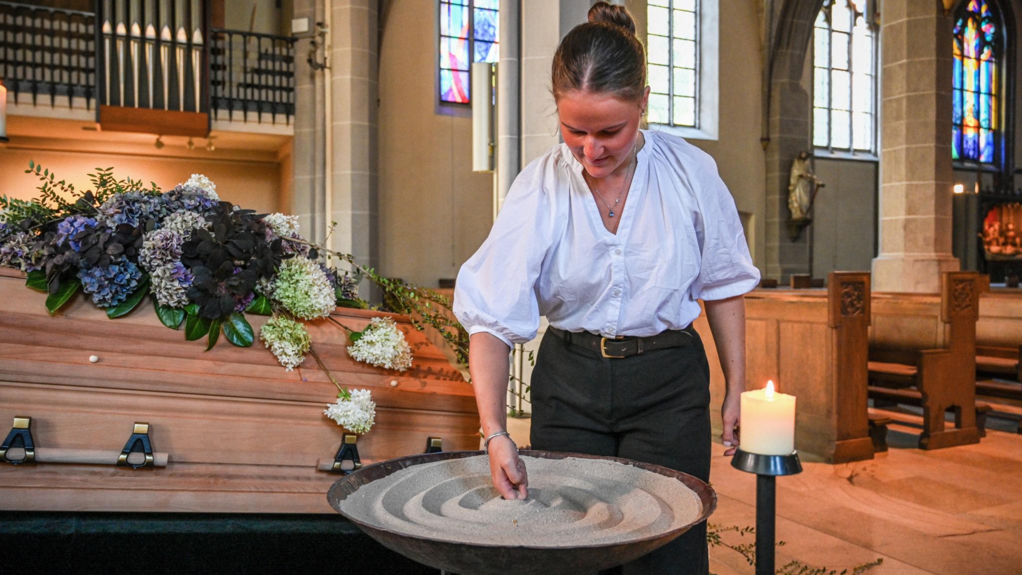Eine Bestatterin gestaltet Sand in einem Gefäß vor einem geschmückten Sarg.