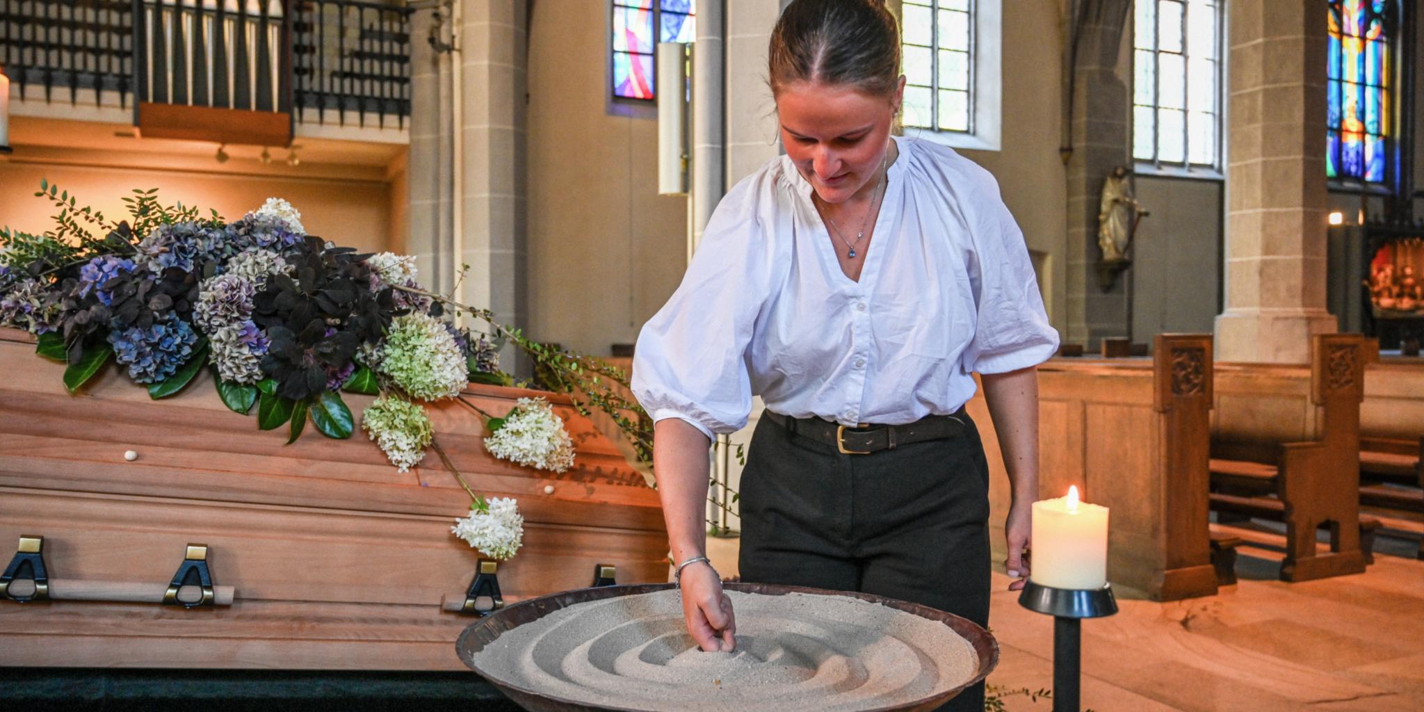 Eine Bestatterin gestaltet Sand in einem Gefäß vor einem geschmückten Sarg.