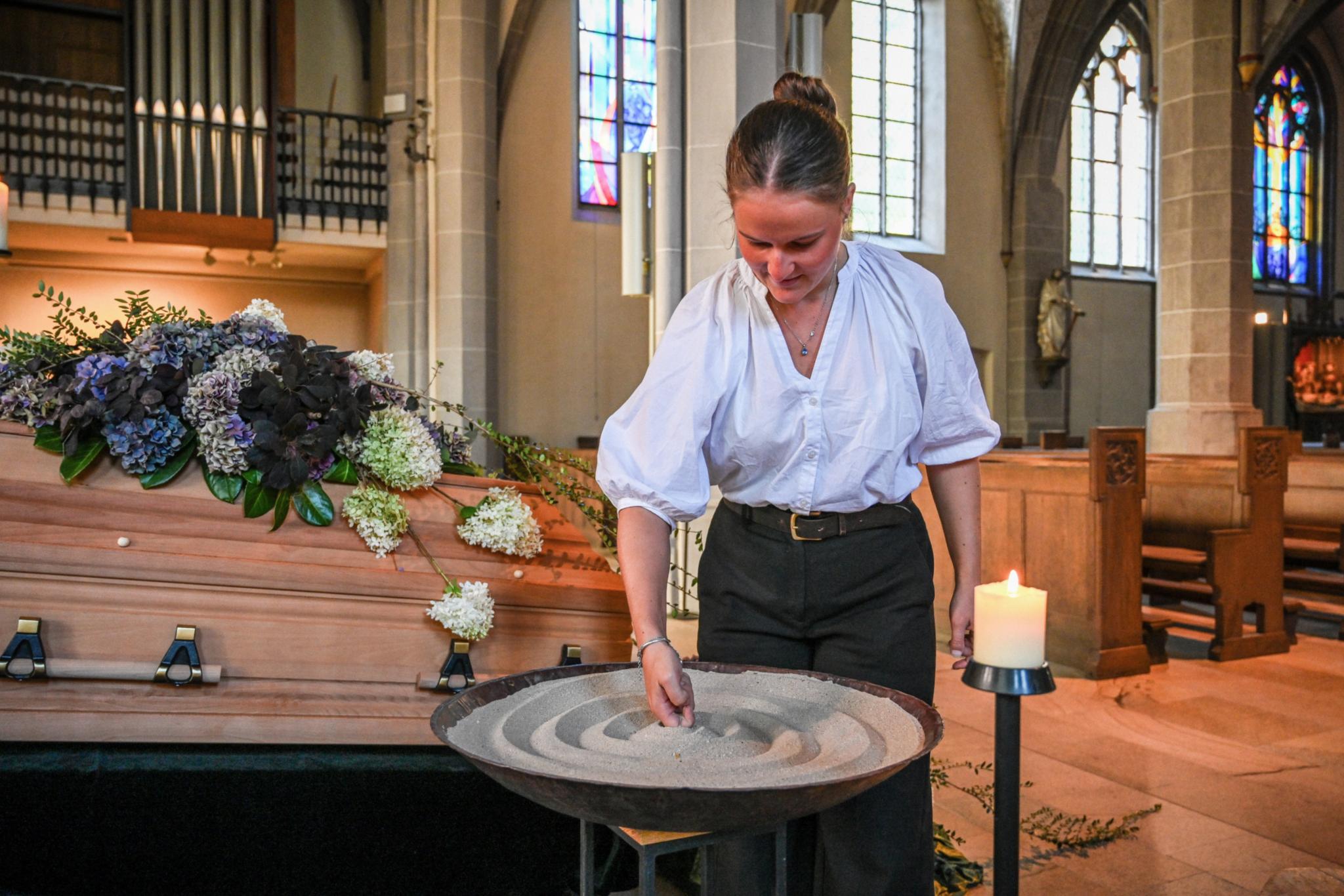 Eine Bestatterin gestaltet Sand in einem Gefäß vor einem geschmückten Sarg.