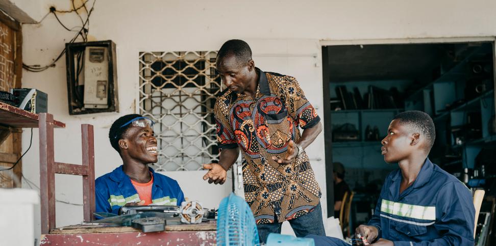 Drei Männer mit dunkler Hautfarbe sitzen an einem Tisch und reparieren etwas
