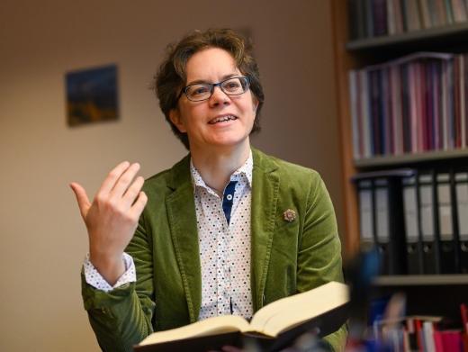 Eine Frau im grünen Blazer hält ein Buch in der Hand und spricht