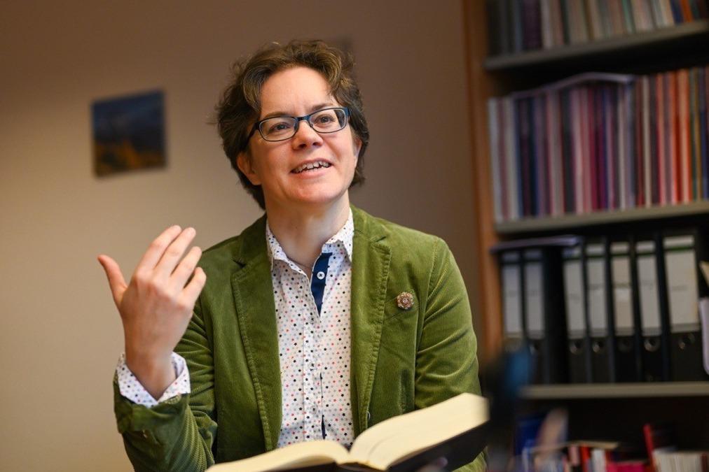 Eine Frau im grünen Blazer hält ein Buch in der Hand und spricht