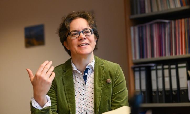 Eine Frau im grünen Blazer hält ein Buch in der Hand und spricht