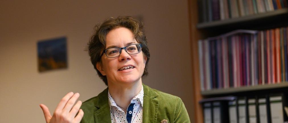 Eine Frau im grünen Blazer hält ein Buch in der Hand und spricht