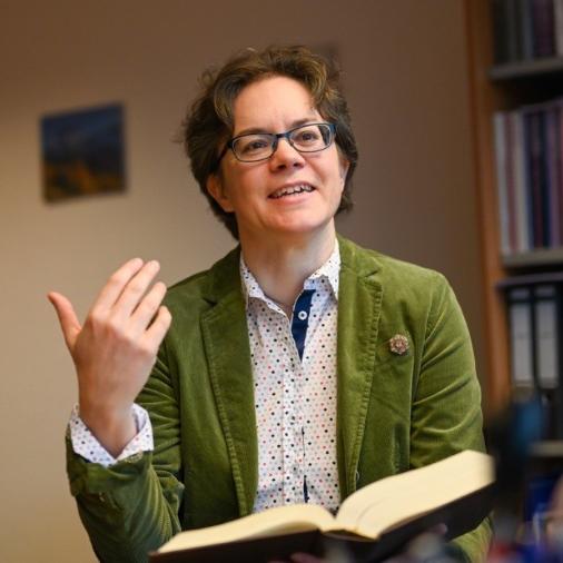 Eine Frau im grünen Blazer hält ein Buch in der Hand und spricht