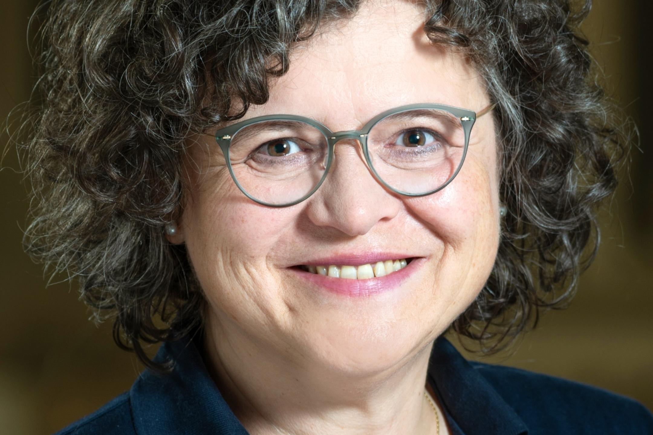 Eine Frau mit dunklen lockigen Haaren und Brille lächelt in die Kamera