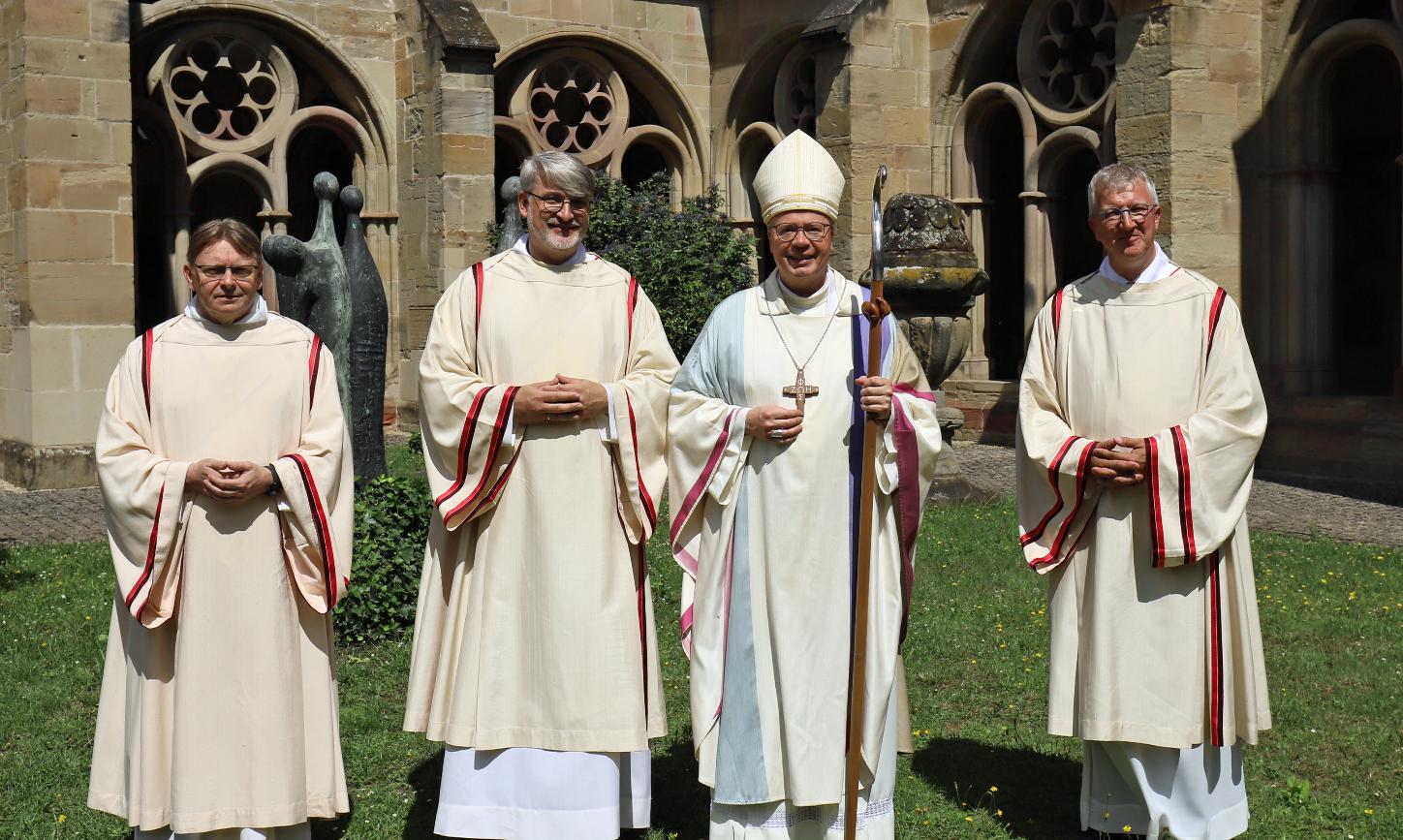 Drei Diakone und ein Bischof in liturgischen Gewändern vor einer Kirche