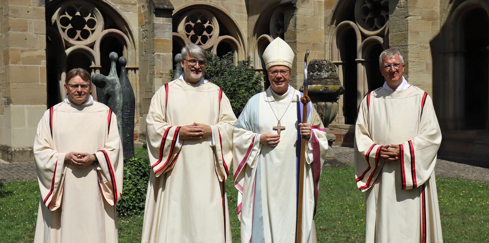 Drei Diakone und ein Bischof in liturgischen Gewändern vor einer Kirche