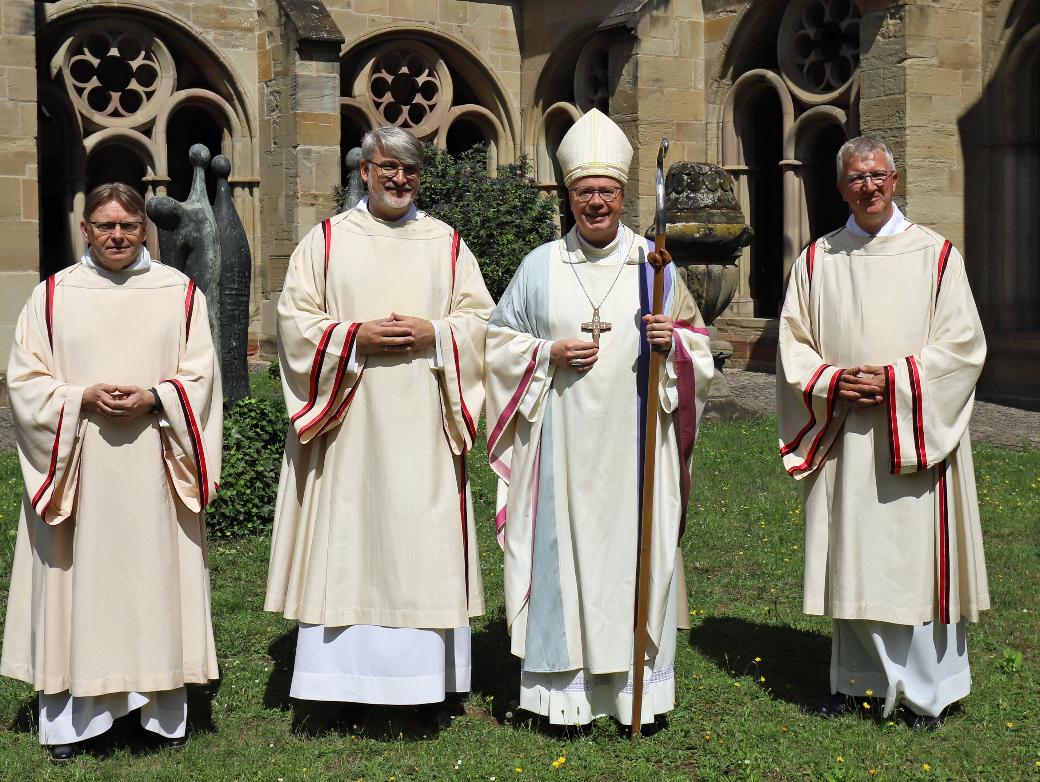 Drei Diakone und ein Bischof in liturgischen Gewändern vor einer Kirche