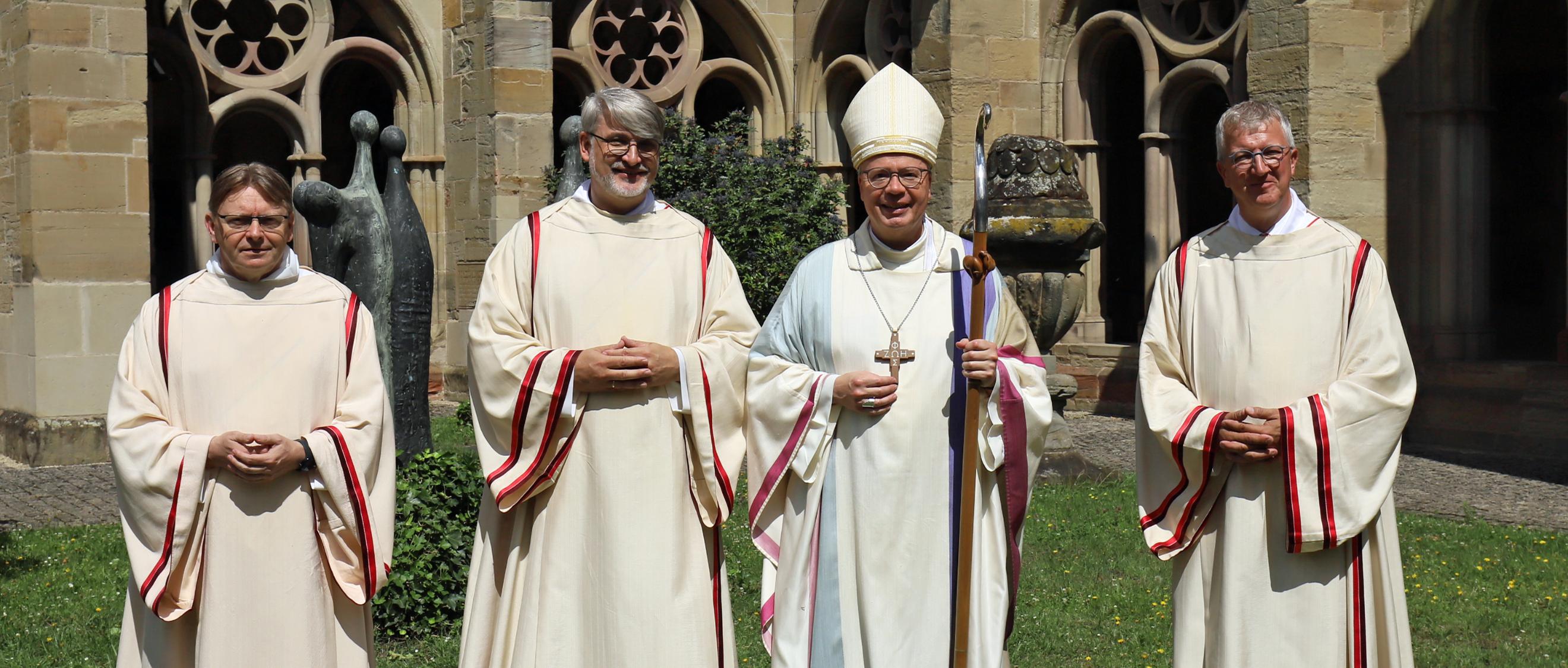 Drei Diakone und ein Bischof in liturgischen Gewändern vor einer Kirche