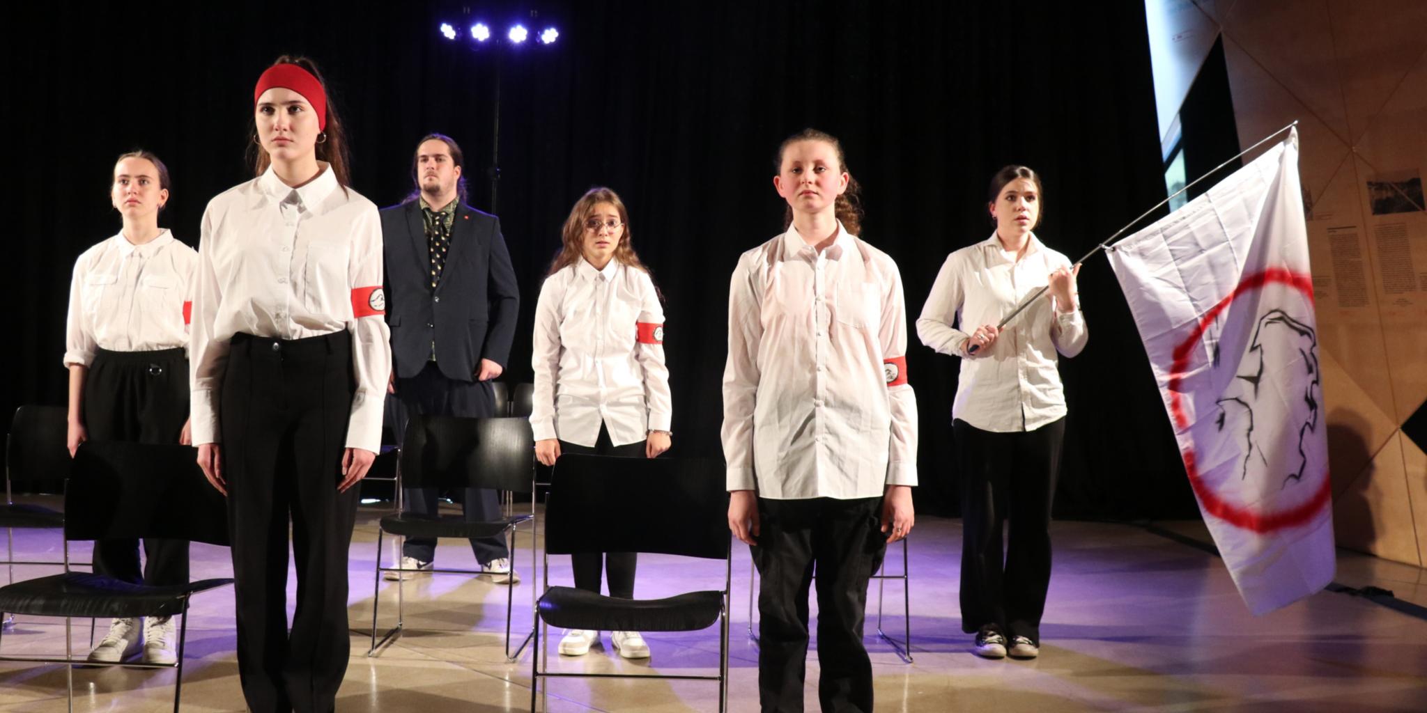 Die Gruppendynamik macht sich auch Eine Schülergruppe mit Lehrer steht 'stramm' neben ihren Stühlen. Die Schüler tragen alle weiße Blusen, schwarze Hosen und ein rotes Armband mit Wellensymbol.