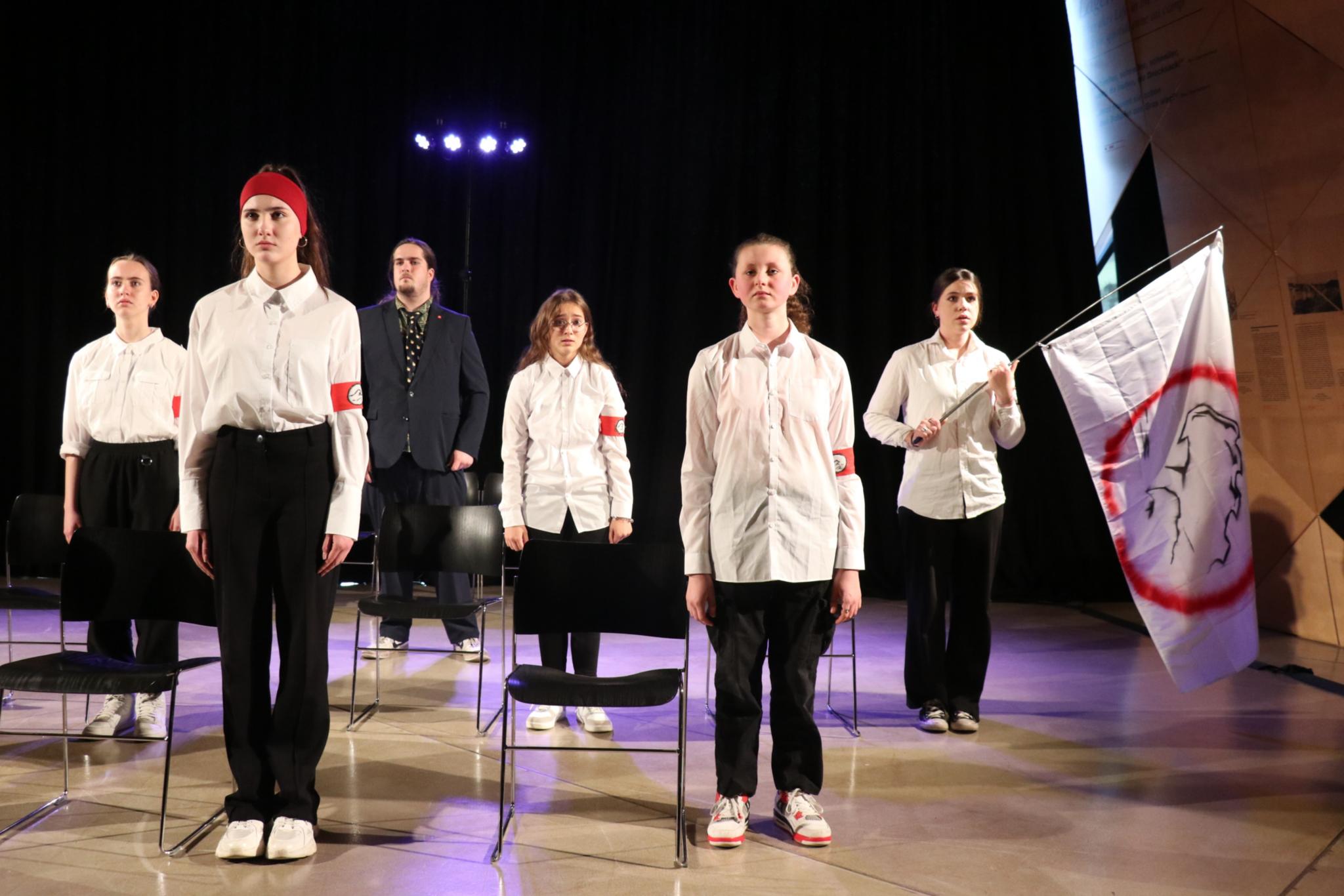 Die Gruppendynamik macht sich auch Eine Schülergruppe mit Lehrer steht 'stramm' neben ihren Stühlen. Die Schüler tragen alle weiße Blusen, schwarze Hosen und ein rotes Armband mit Wellensymbol.