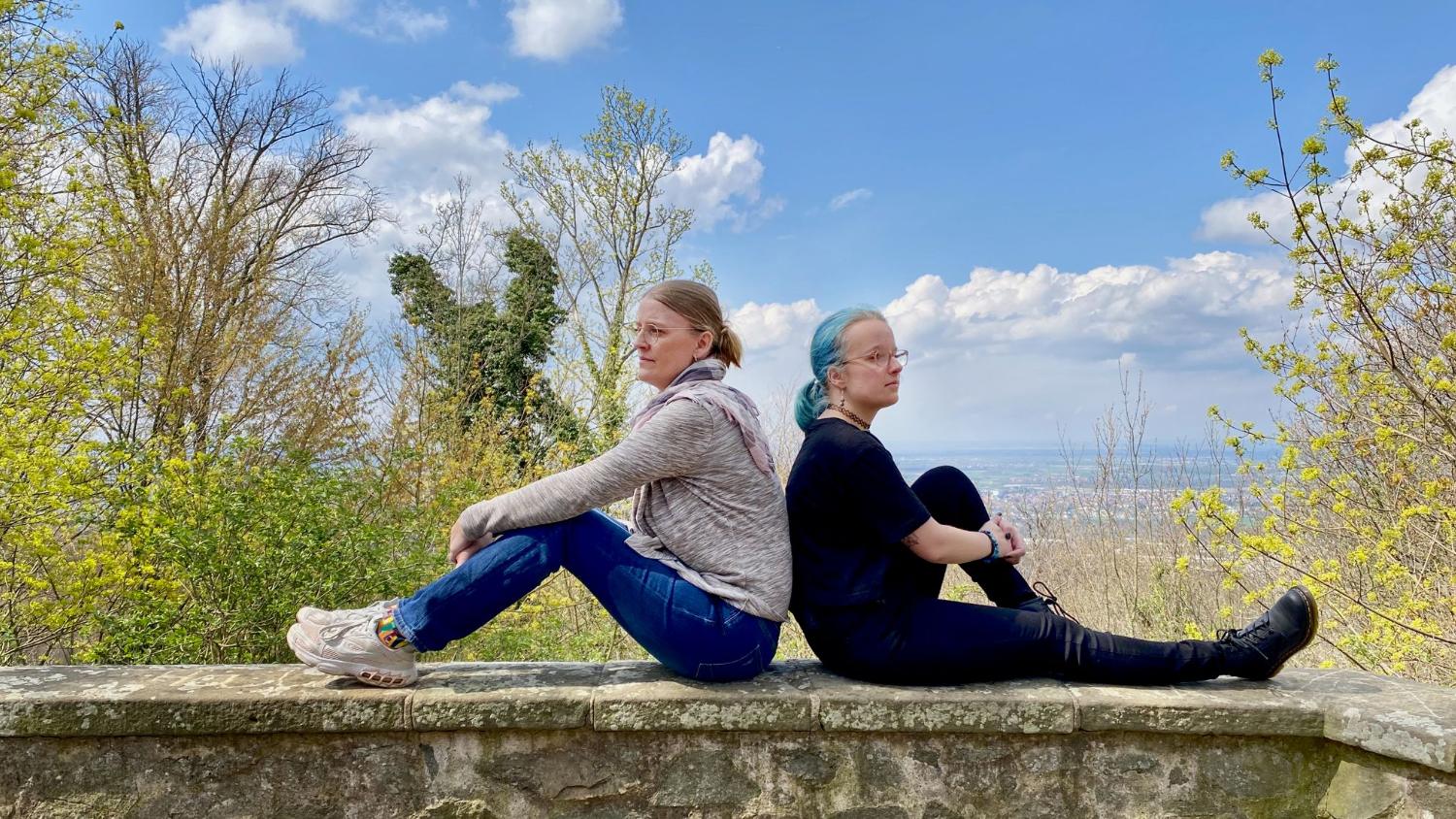 Zwei Frauen sitzen Rücken an Rücken auf einer Mauer.