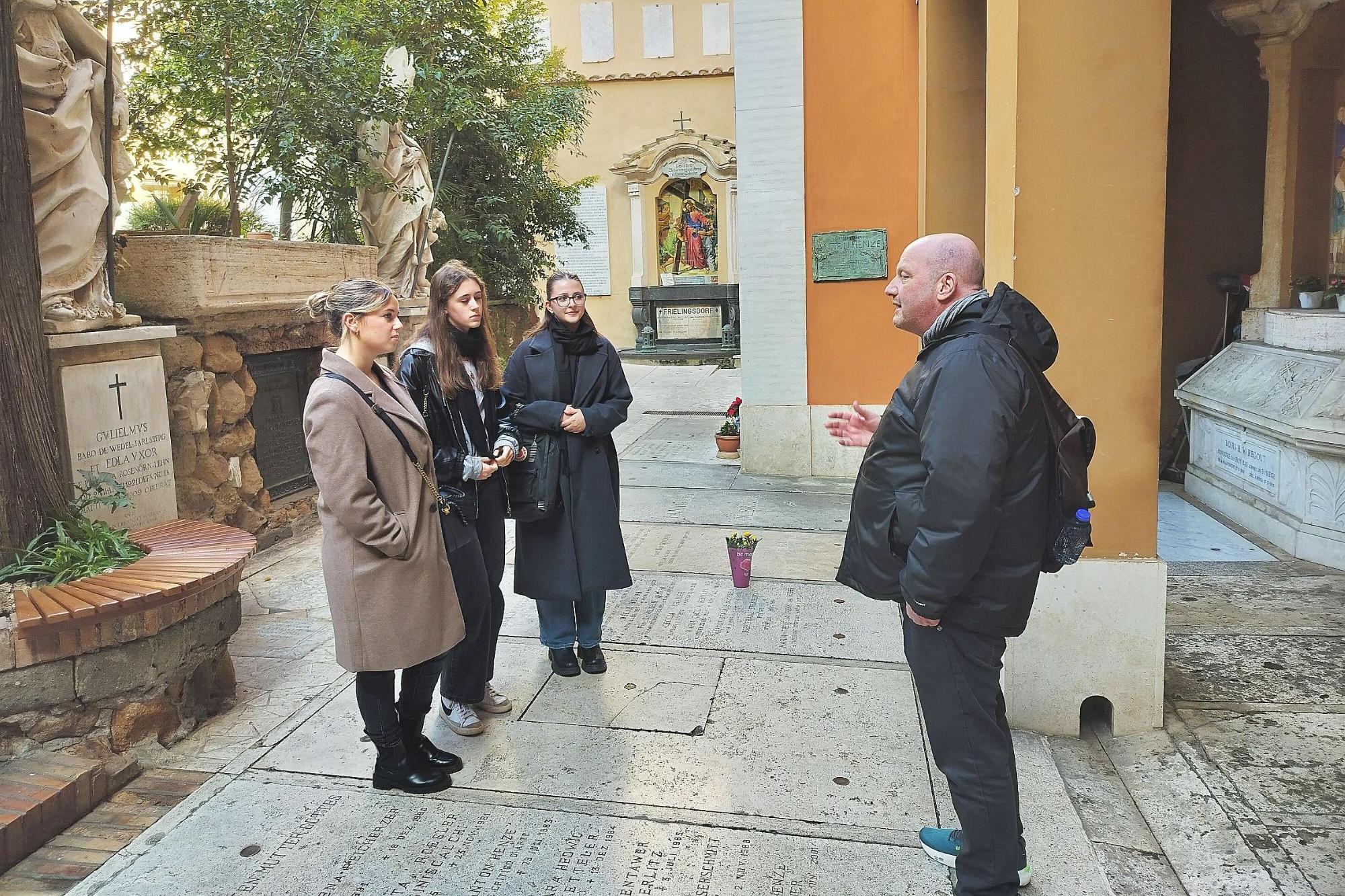 Drei junge Frauen stehen mit einem Mann auf einem Friedhof und hören seinen Erklärungen zu