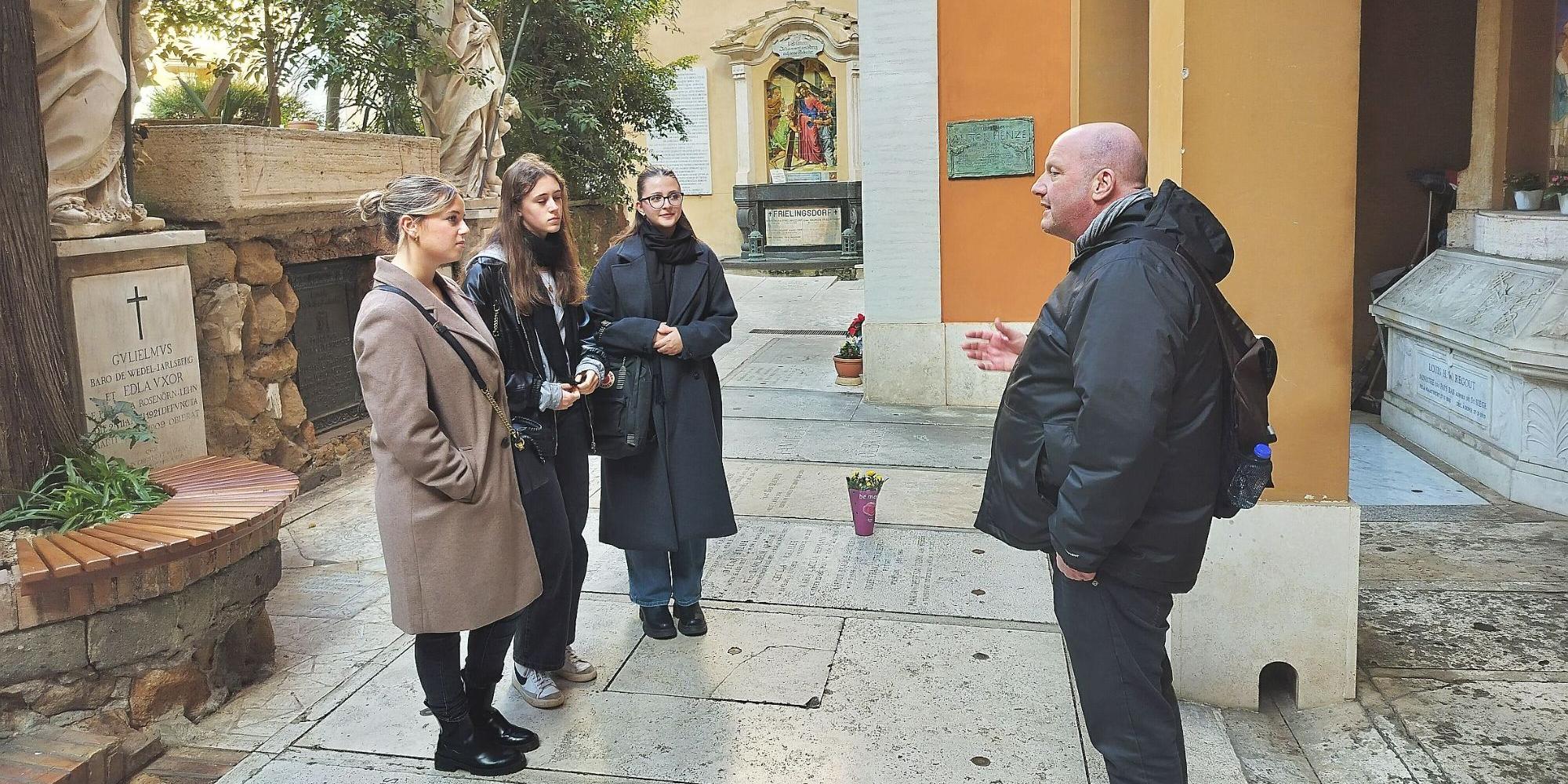 Drei junge Frauen stehen mit einem Mann auf einem Friedhof und hören seinen Erklärungen zu