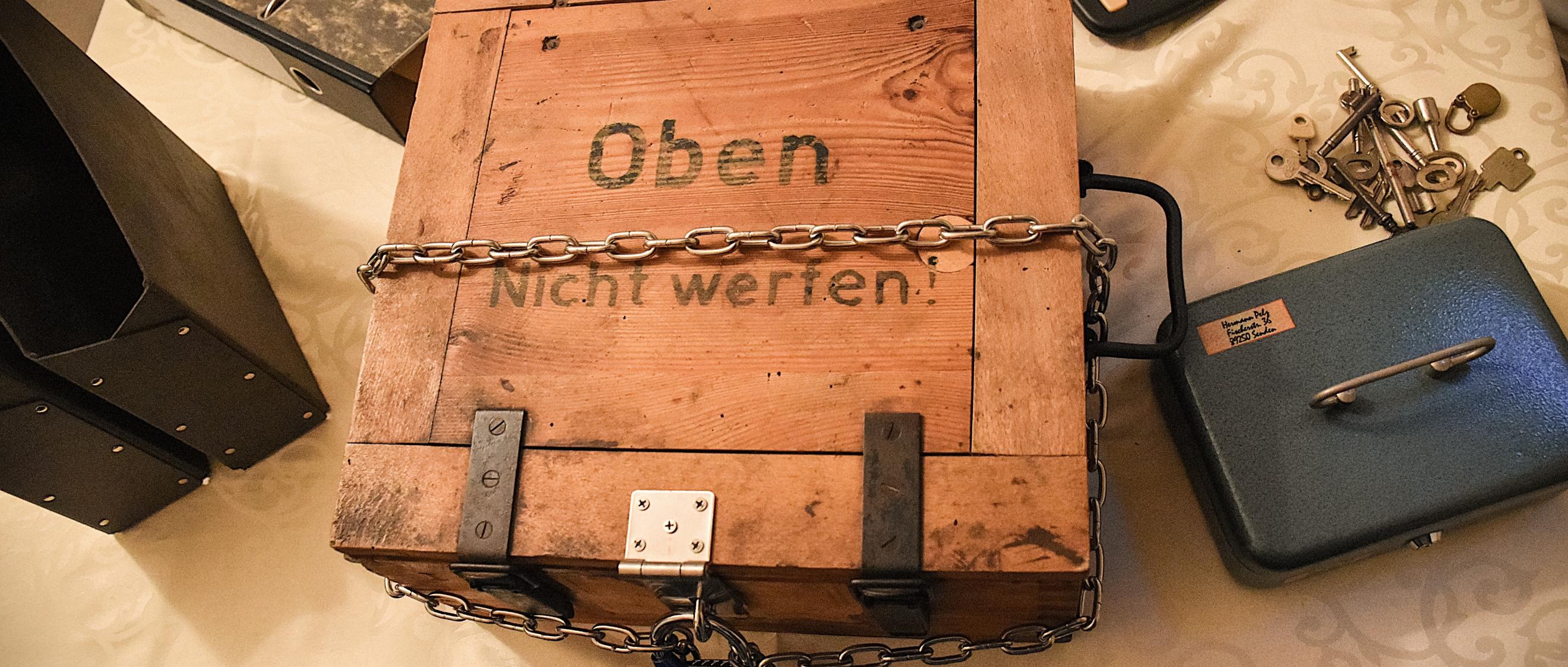 Kiste, Ordner, Taschenrechner, Schlüssel und Kasse verteilt auf einem Tisch sind Bestandteile des Rätsels.