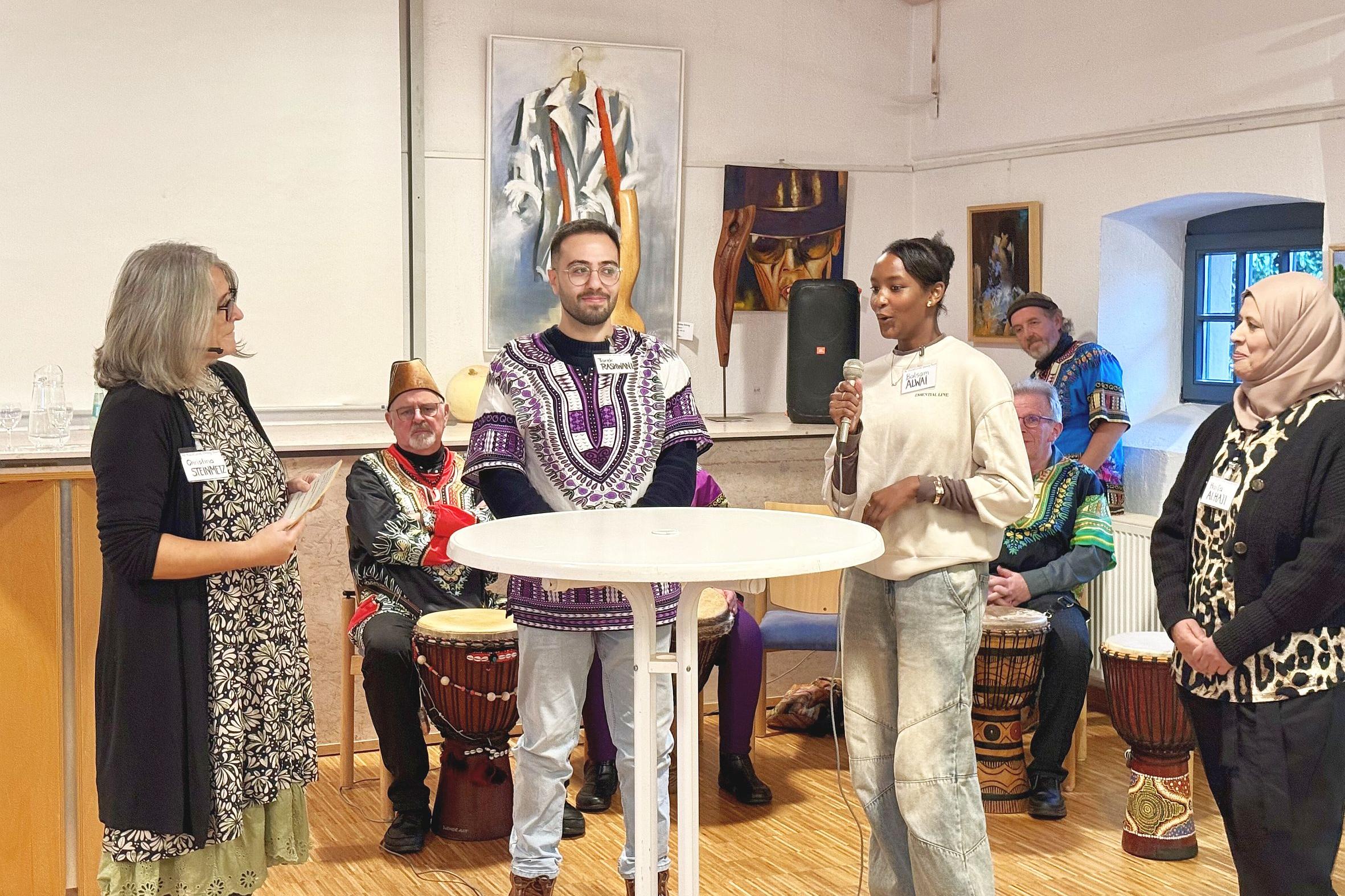 Vier Personen an einem Stehtisch, eine davon spricht in ein Mikrofon. Im Hintergrund sitzen Trommler in Tracht.