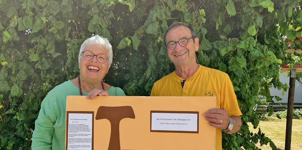 Eine Frau in Grünem T-Shurt und ein Mann in gelbem T-Shirt halten ein Plakat mit dem Franziskanischen Tau in die Kamera