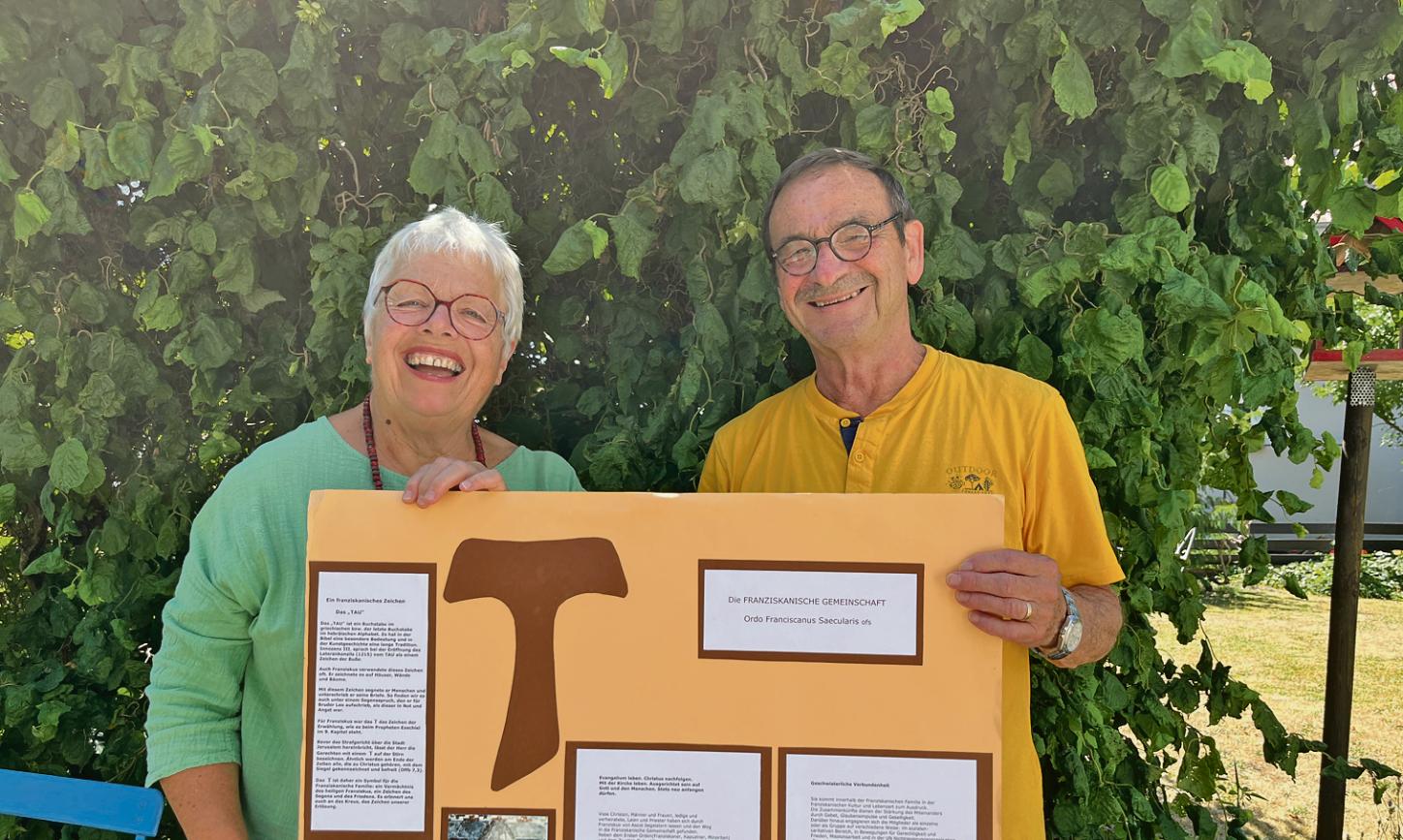 Eine Frau in Grünem T-Shurt und ein Mann in gelbem T-Shirt halten ein Plakat mit dem Franziskanischen Tau in die Kamera