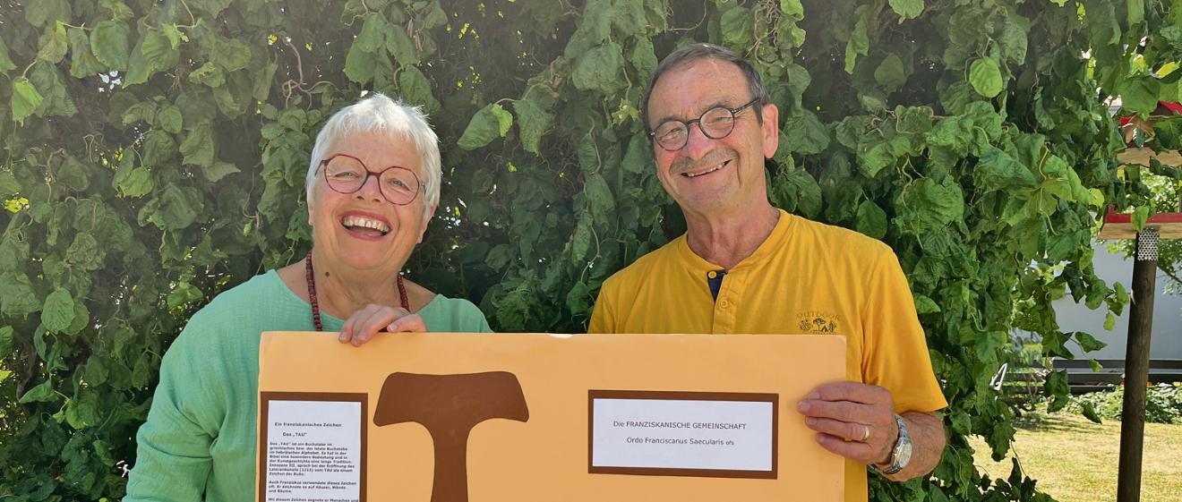 Eine Frau in Grünem T-Shurt und ein Mann in gelbem T-Shirt halten ein Plakat mit dem Franziskanischen Tau in die Kamera
