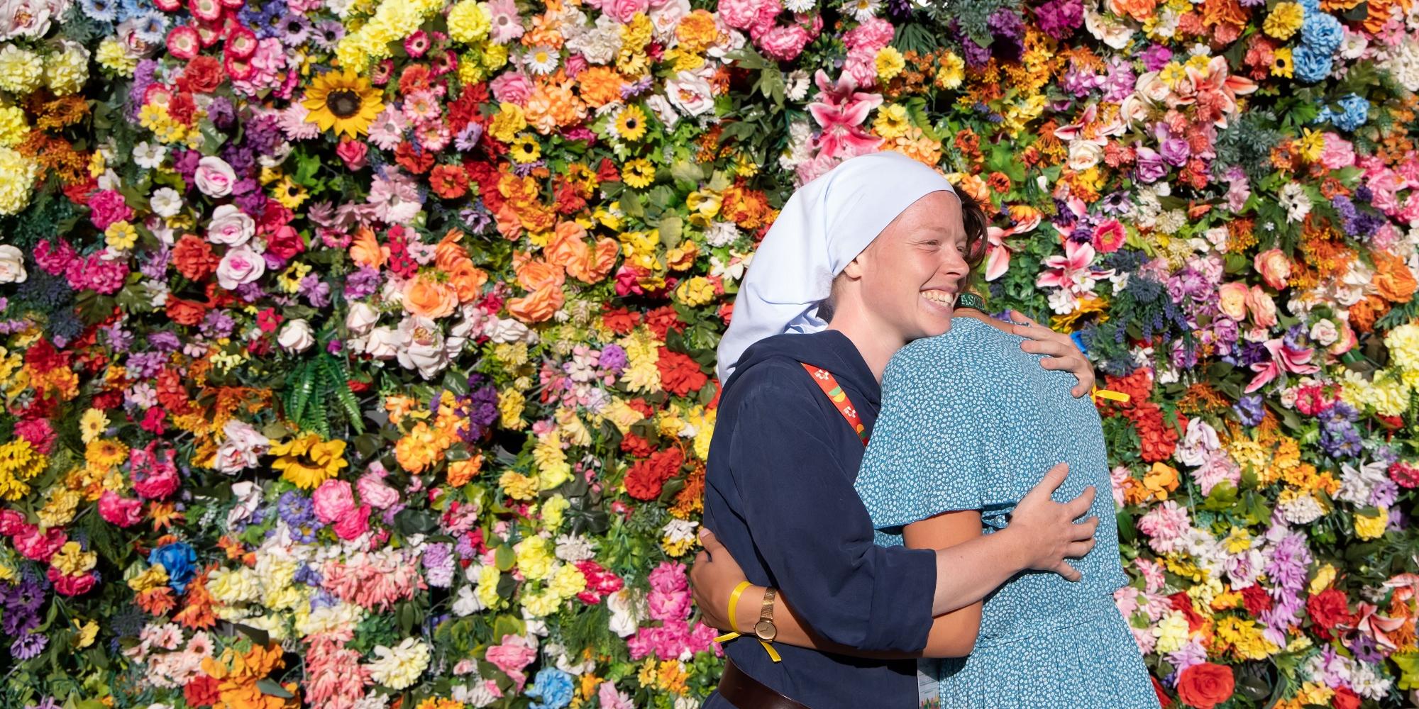 Zwei Frauen umarmen sich vor einer Wand aus Blumen.