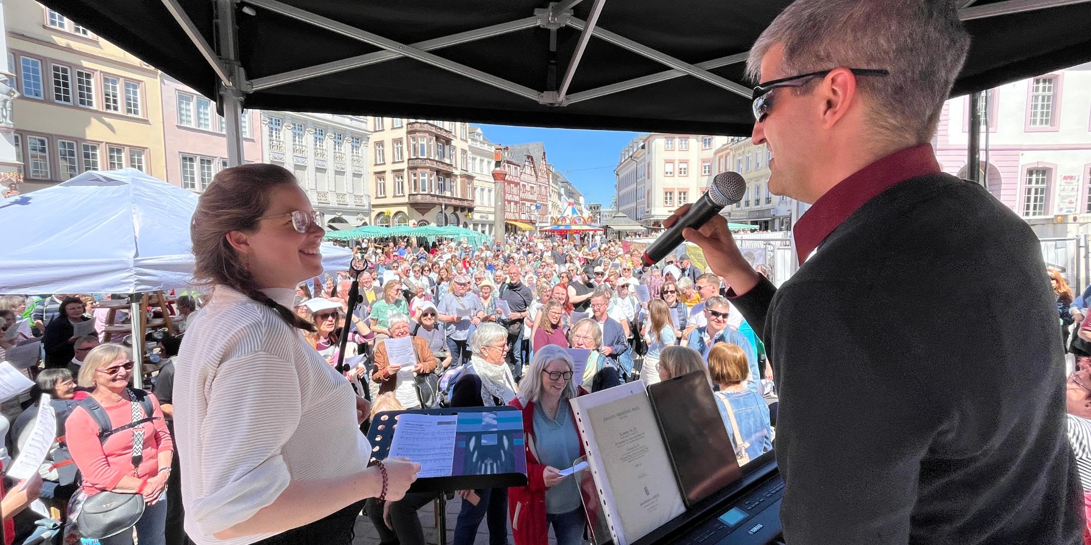Ein Mann am Klavier und eine Frau stehen auf einer Bühne mit Blick auf die Menge davor