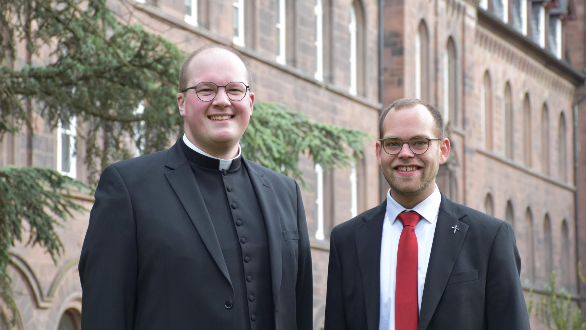 Zwei Männer vor einem rötlichen Gebäude lächeln in die Kamera. Einer trägt einen priesterkragen, einer eine rote Krawatte