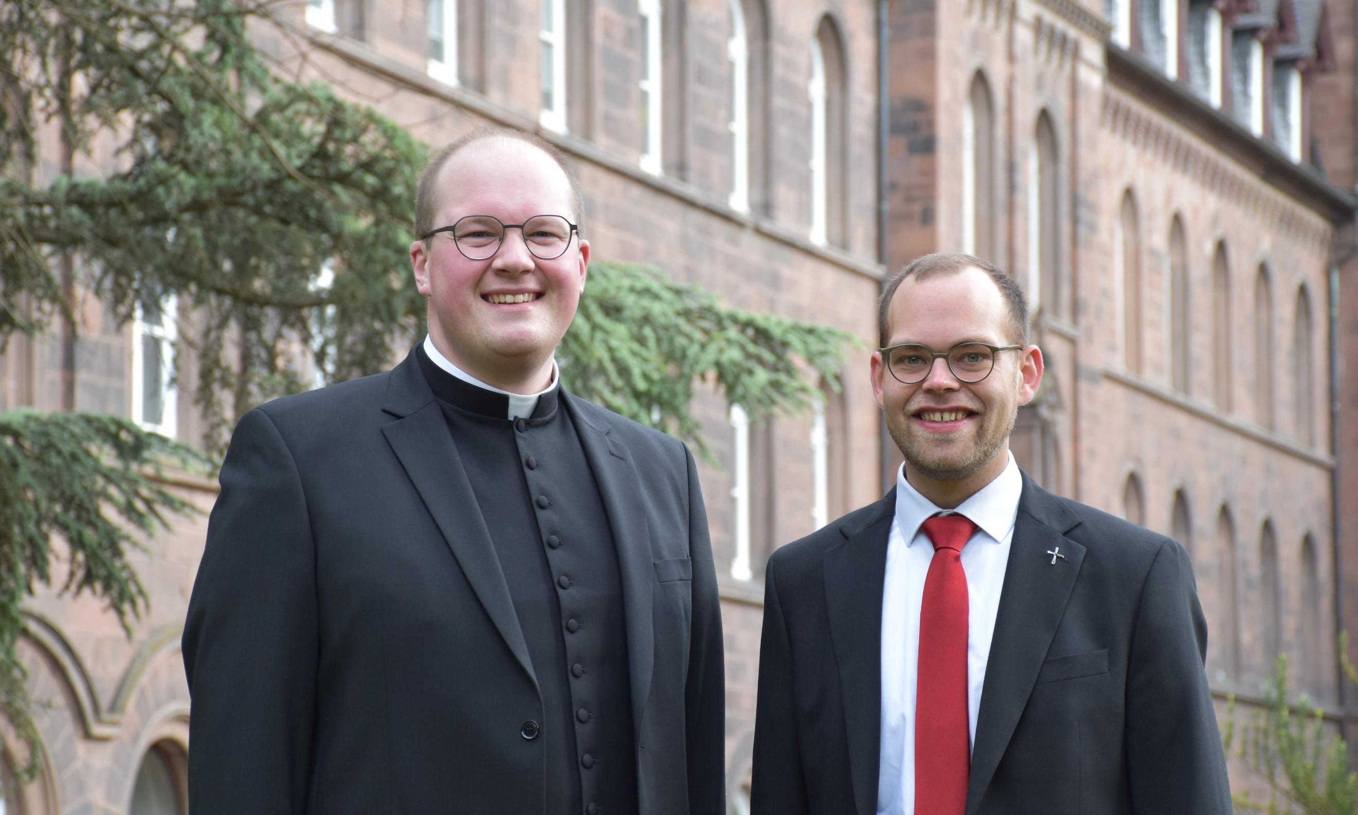 Zwei Männer vor einem rötlichen Gebäude lächeln in die Kamera. Einer trägt einen priesterkragen, einer eine rote Krawatte