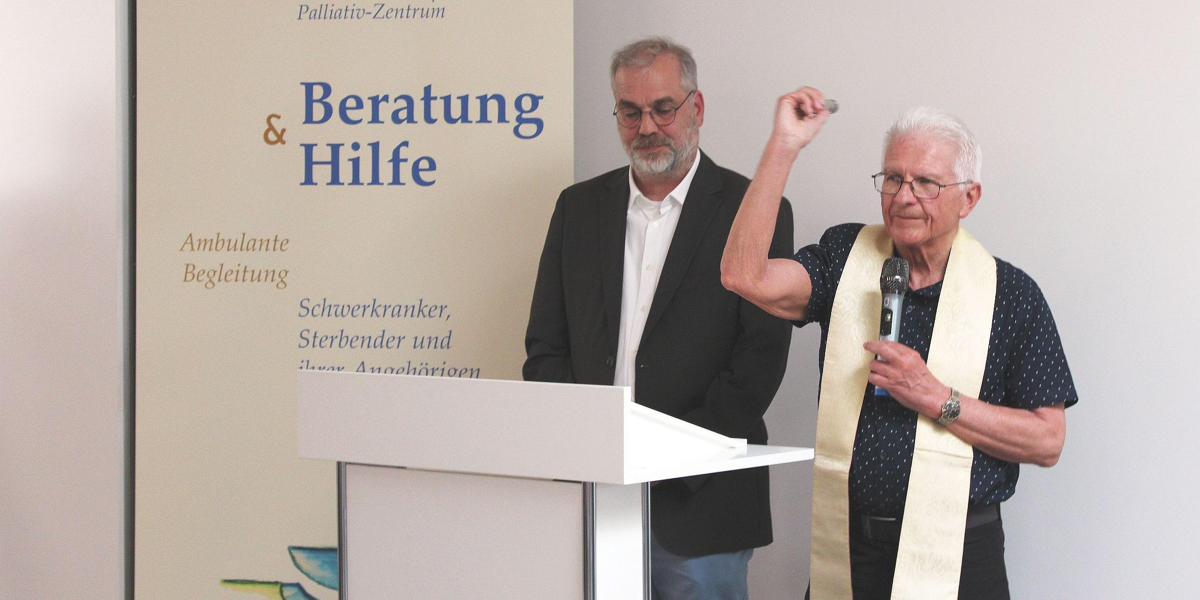 Zwei Männer hinter einem Rednerpult, einer segnet mit Weihwasser