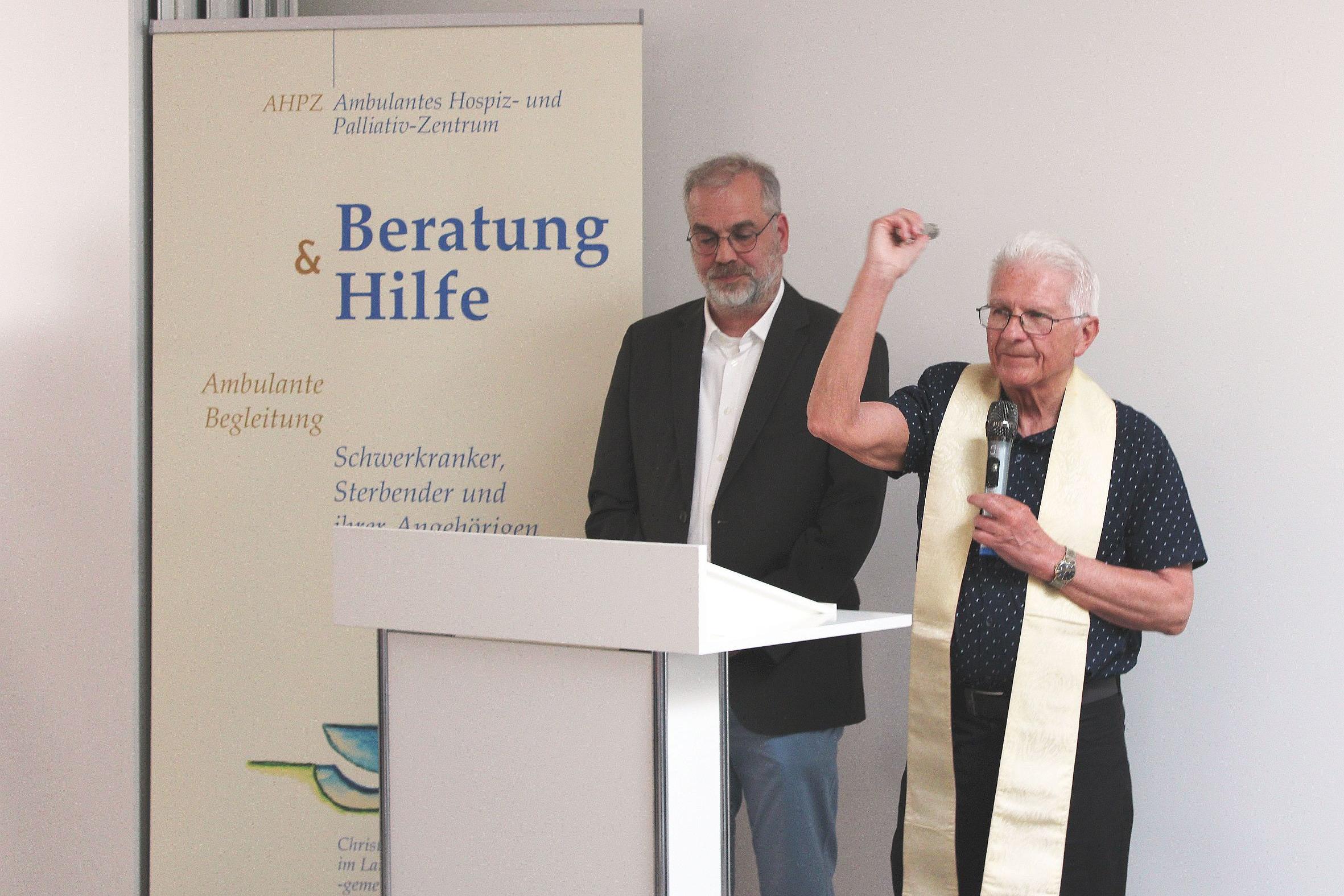 Zwei Männer hinter einem Rednerpult, einer segnet mit Weihwasser