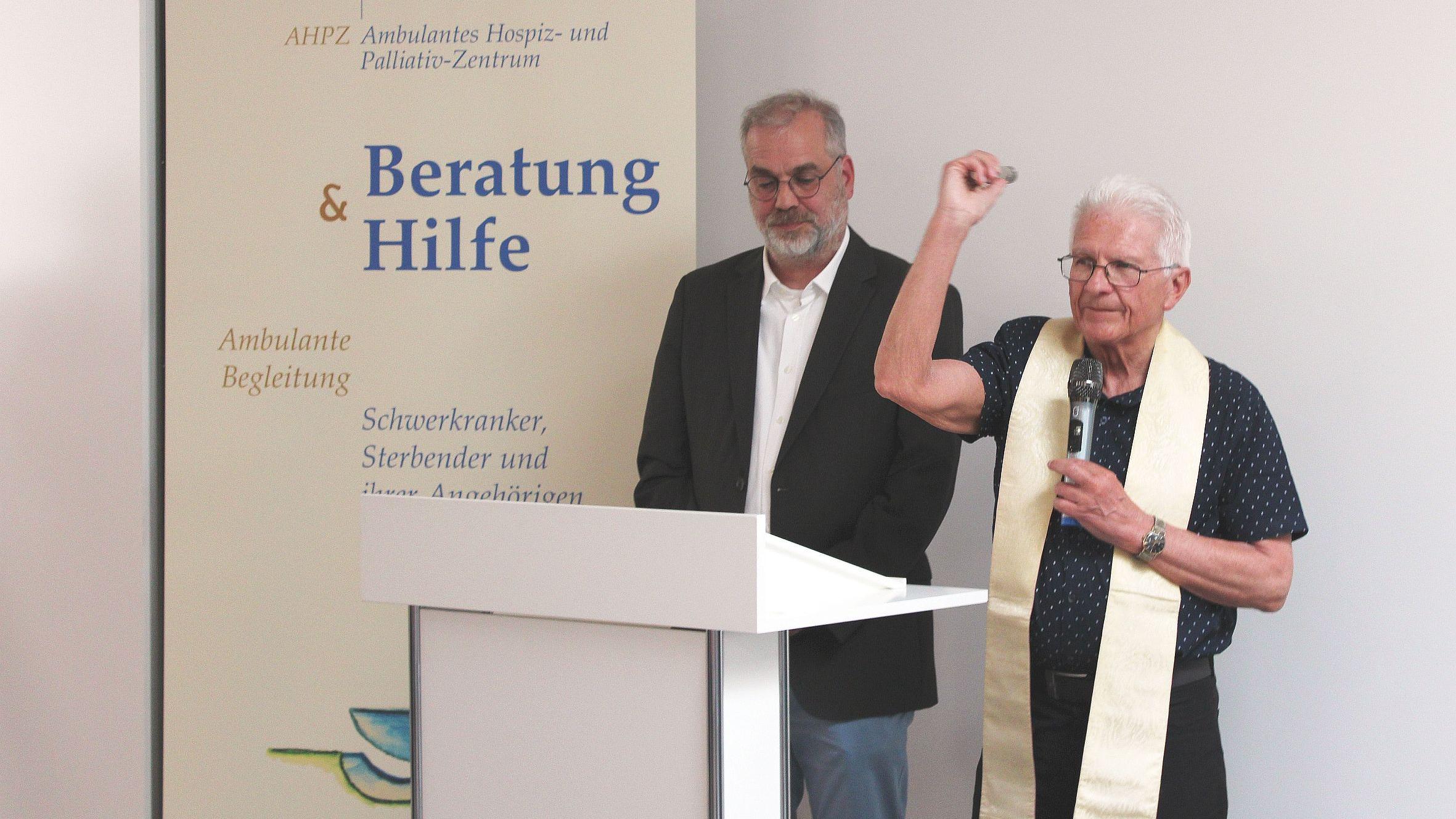 Zwei Männer hinter einem Rednerpult, einer segnet mit Weihwasser