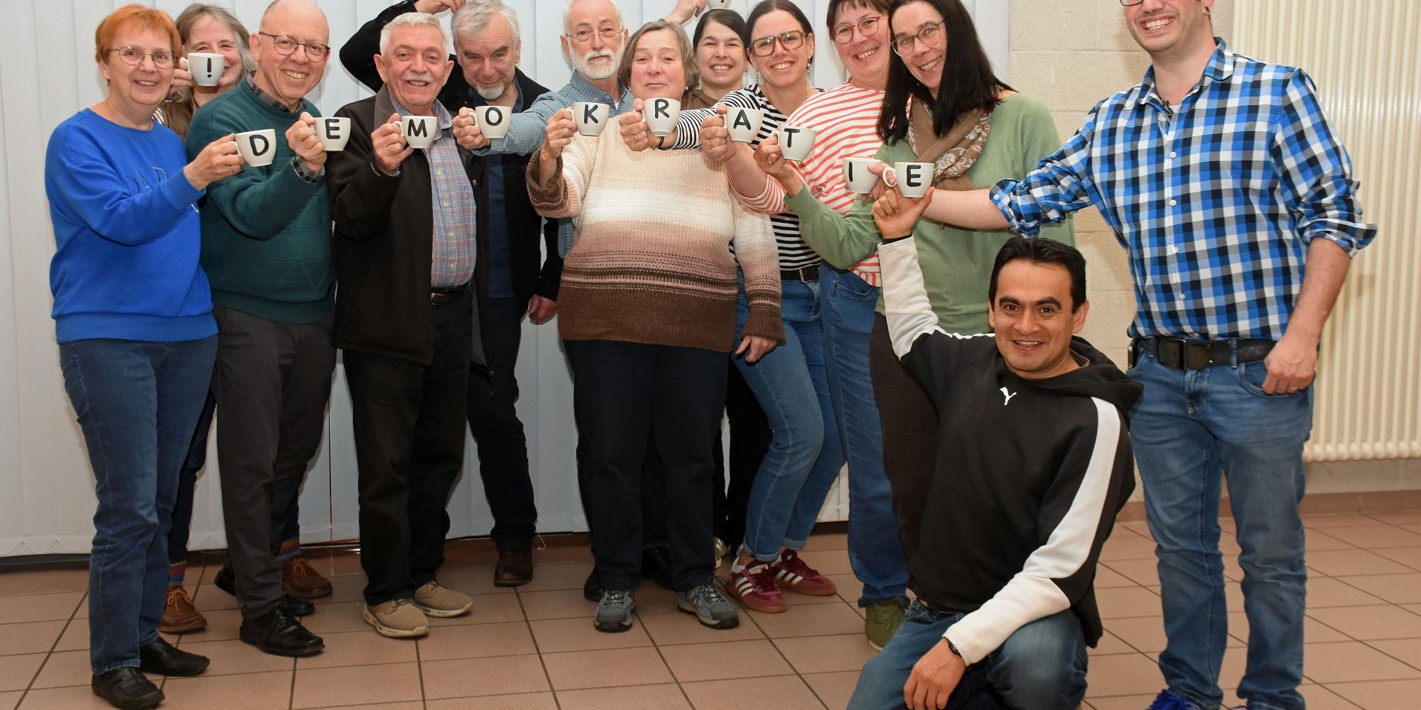 Die Mitglieder des Pfarrgemeinderates halten mit Buchstaben bedruckte Tassen, die zusammen das Wort Demokratie ergeben.