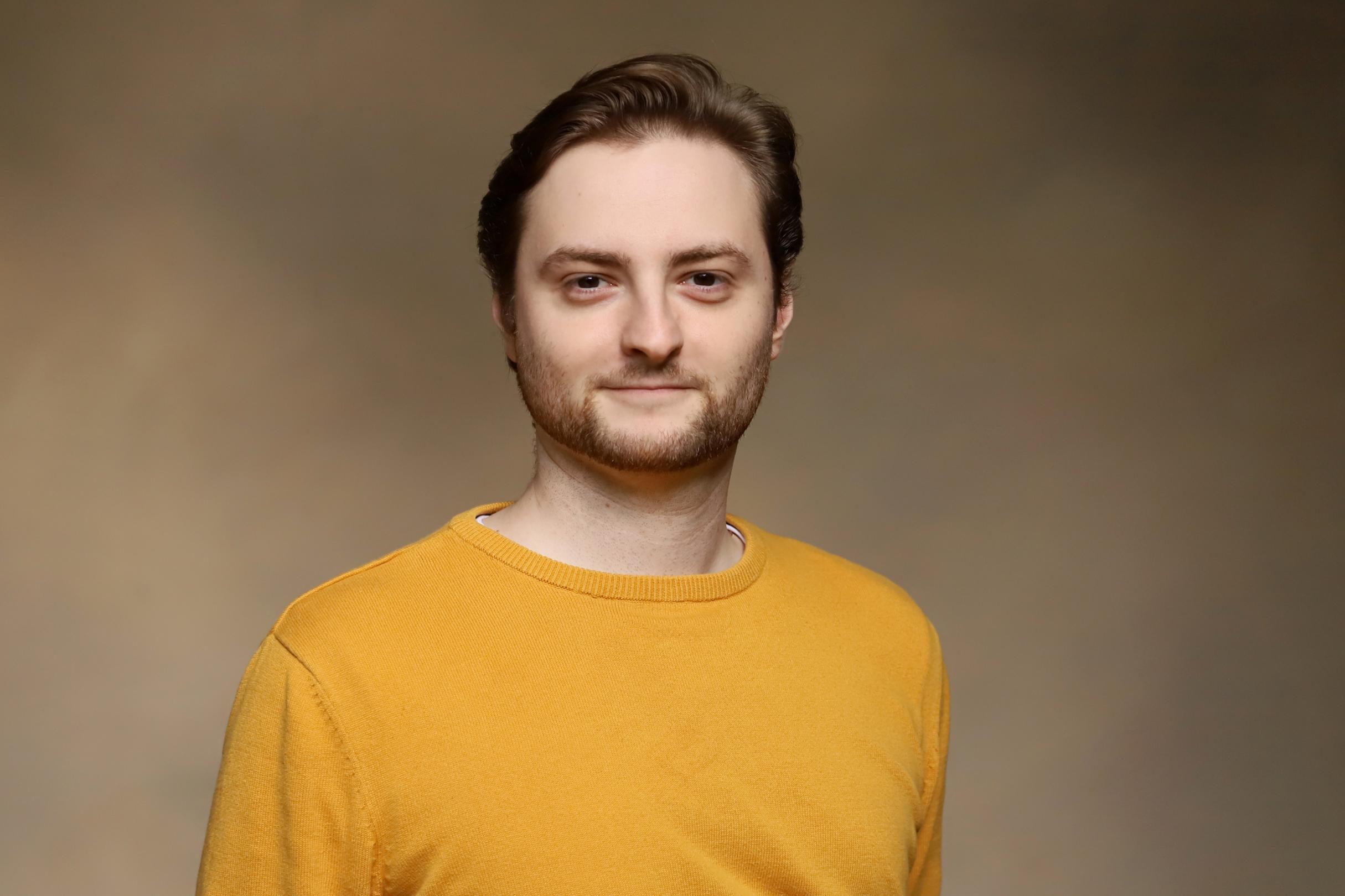 Ein junger Mann im senfgelben Pullover lächelt in die Kamera