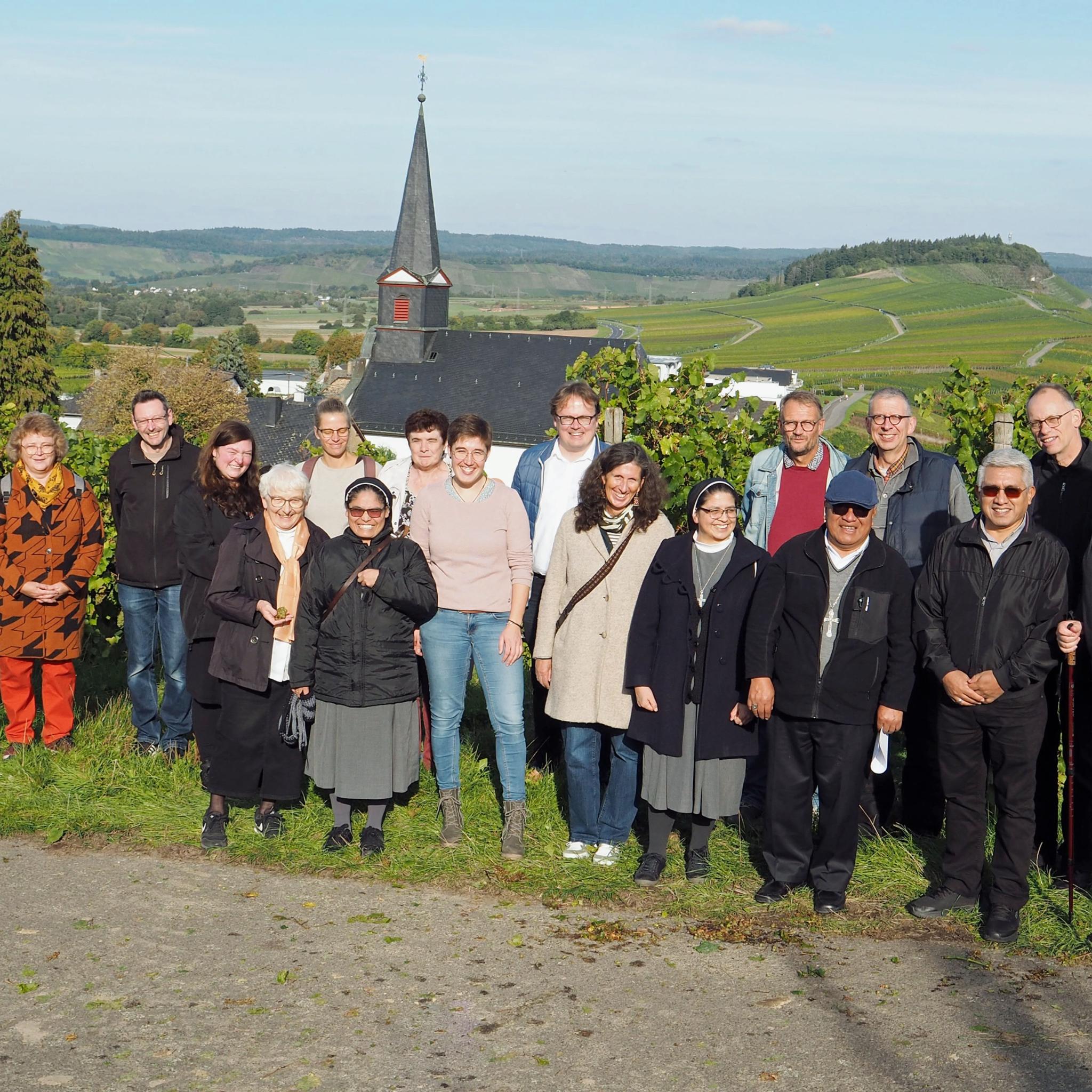 Eine Gruppe Menschen lacht vor einem Panorama mit einer Kirche in die Kamera