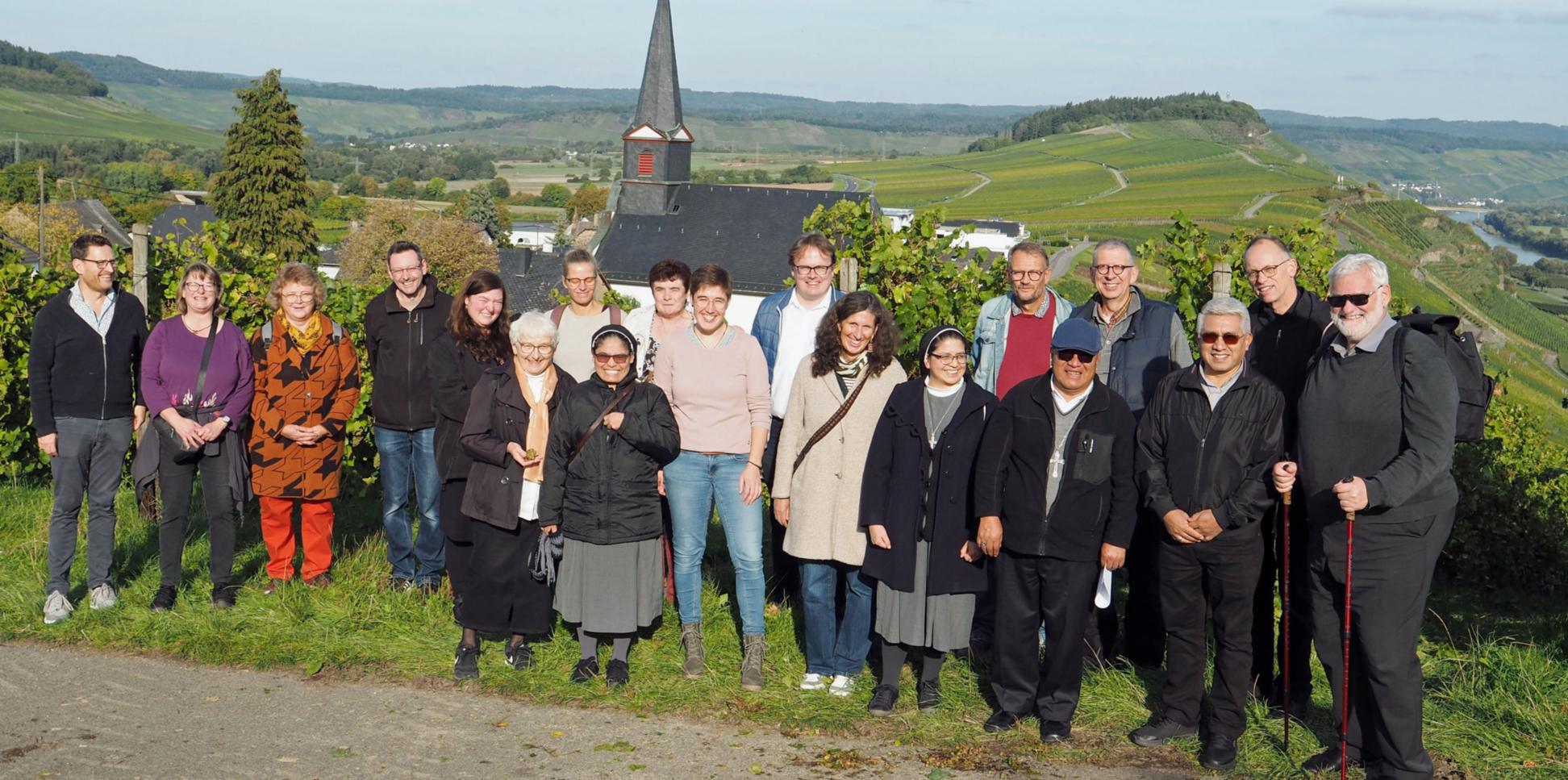 Eine Gruppe Menschen lacht vor einem Panorama mit einer Kirche in die Kamera