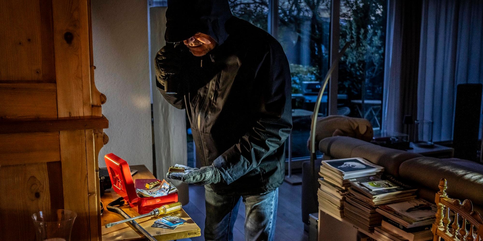 Ein dunkel gekleideter Mann mit Kapuze durchsucht mit einer Taschenlampe eine aufgebrochene Geldkassette.